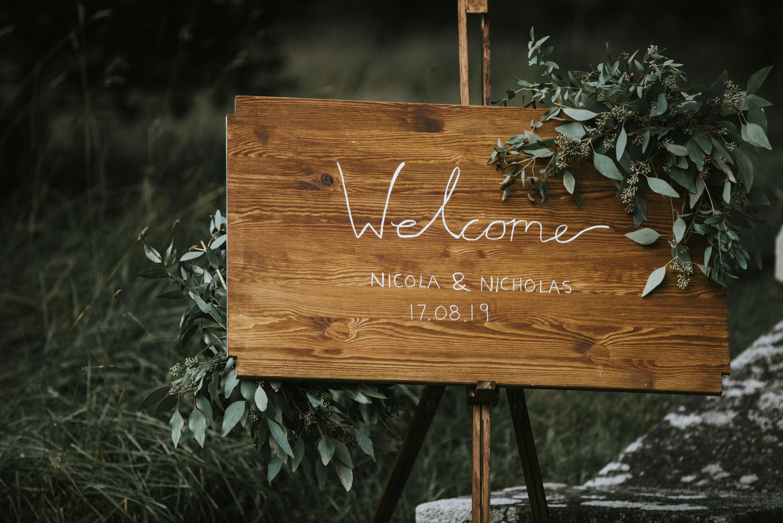 Wooden welcome sign decorated with green foliage, displaying text: "Welcome, Nicola & Nicholas, 17.08.19."