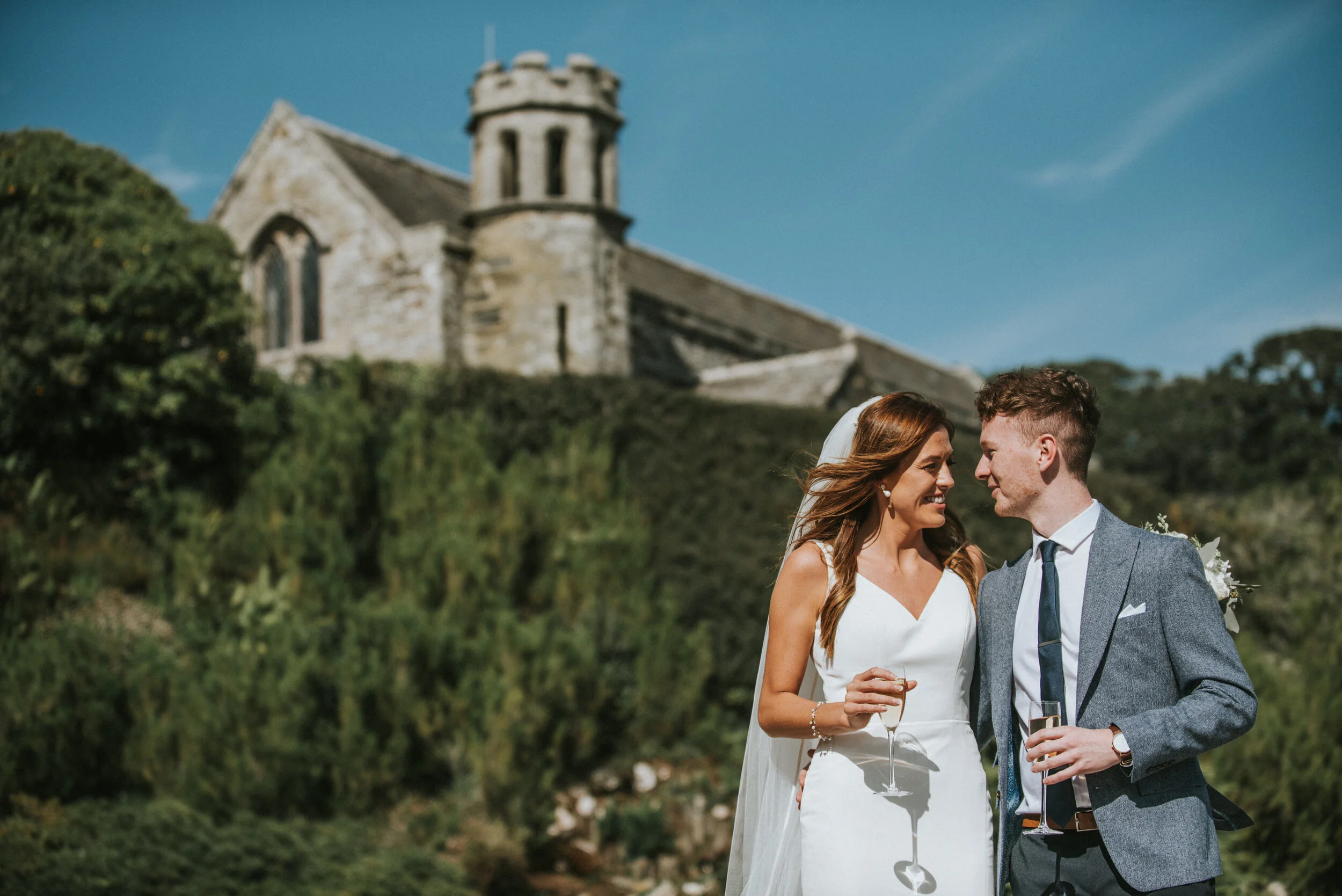 A bride and groom smiling and looking at each other outdoors on their wedding day, holding glasses of champagne, with a historic stone church and green landscape in the background.