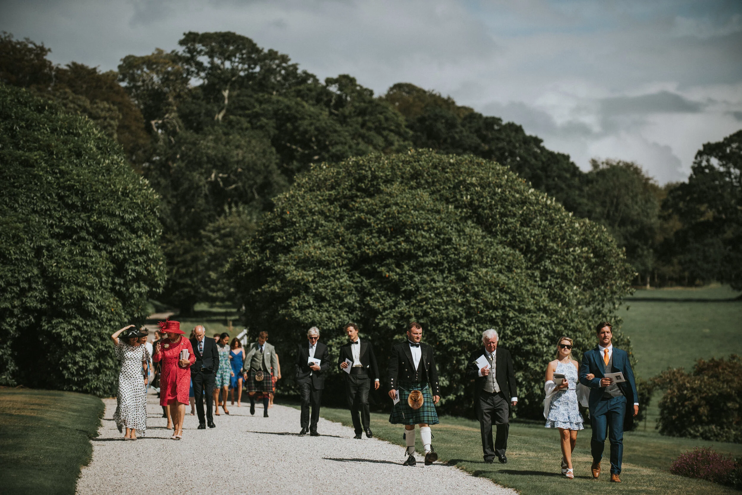 A group of people in formal attire walking on a gravel path through a lush green park, with large trees and a cloudy sky in the background.