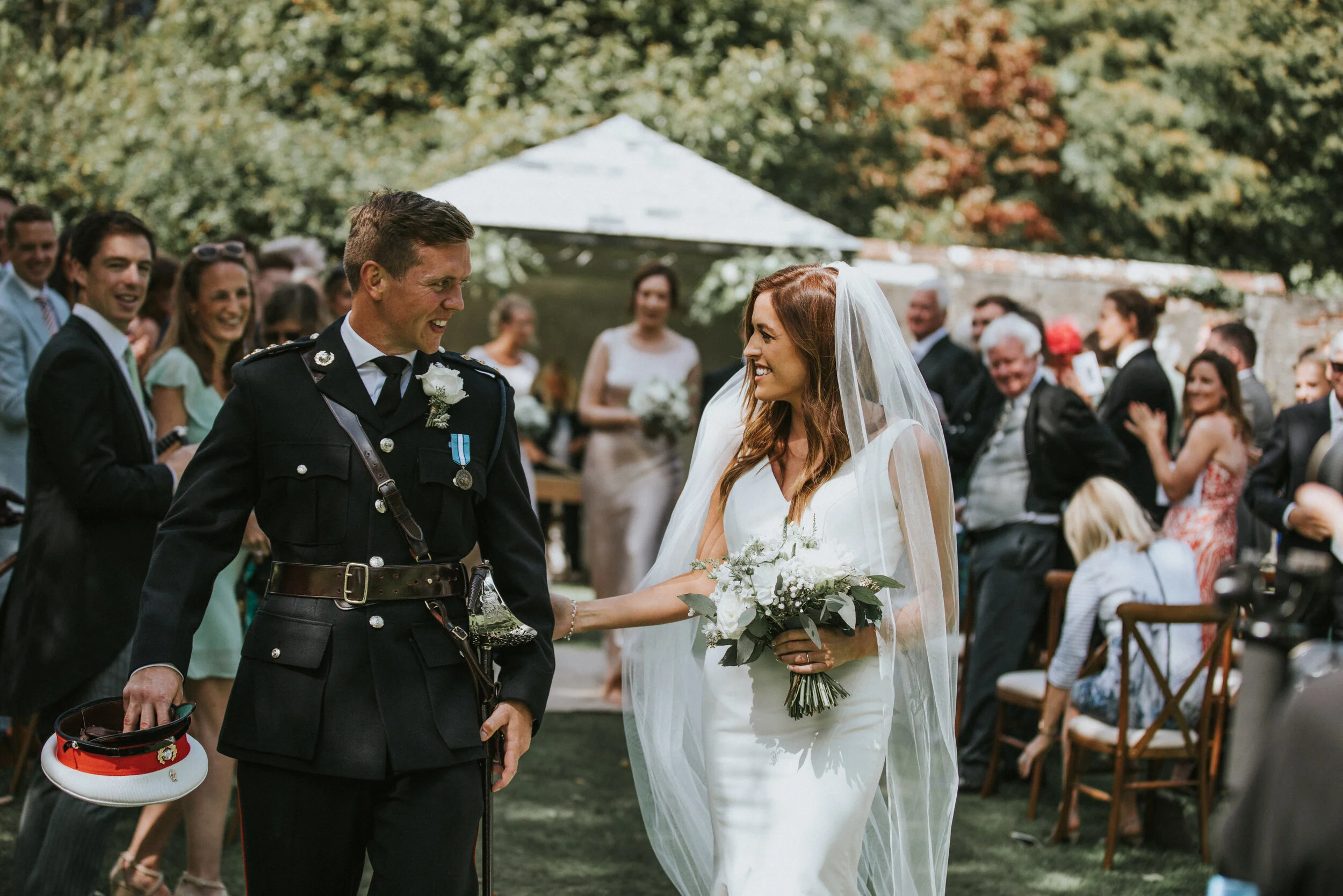 A bride and groom smiling at each other during a wedding ceremony outdoors with guests in the background.