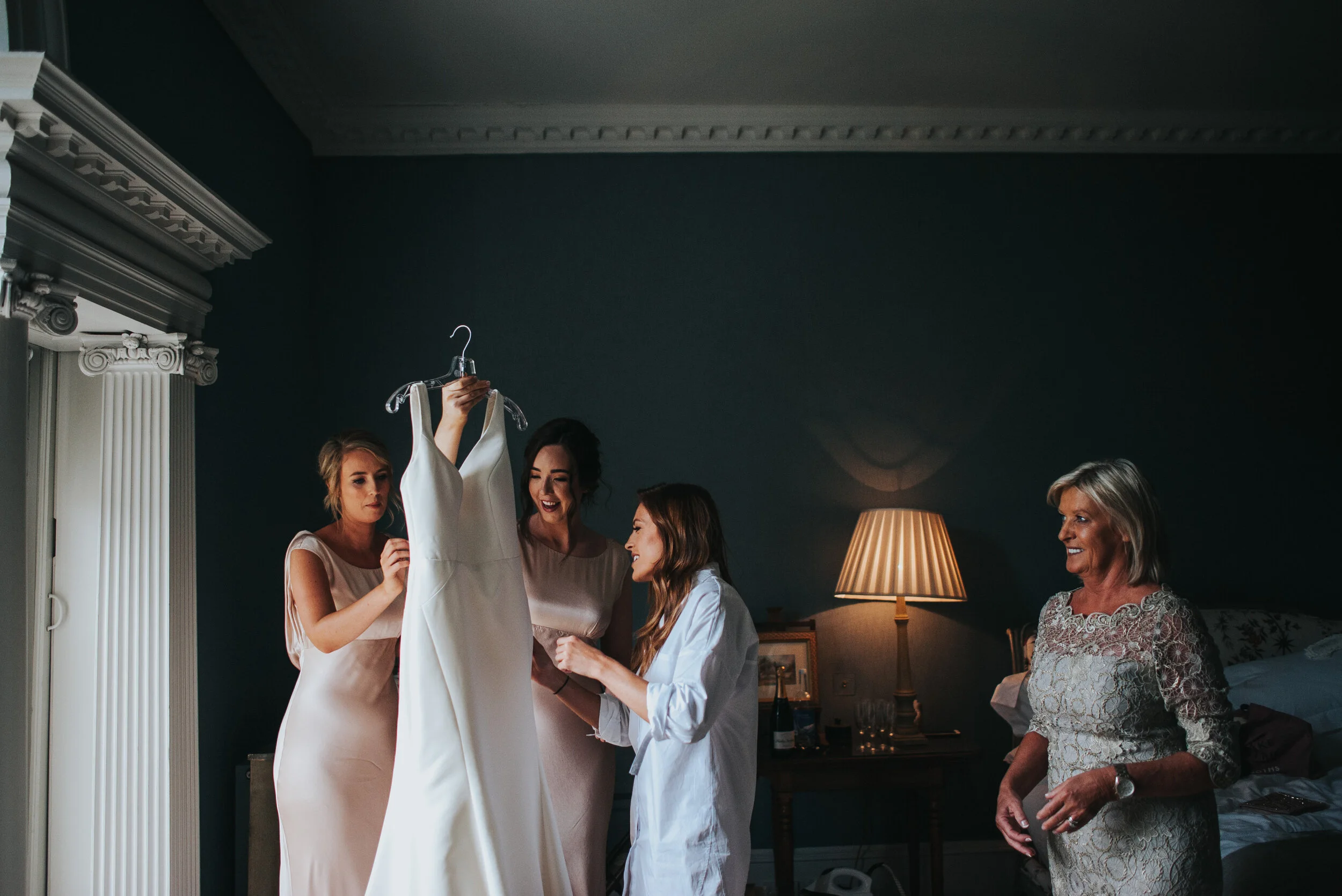 Bridesmaids and an older woman look at a wedding dress hanging on a hanger in a dimly lit room with blue walls and a lamp on a nightstand.