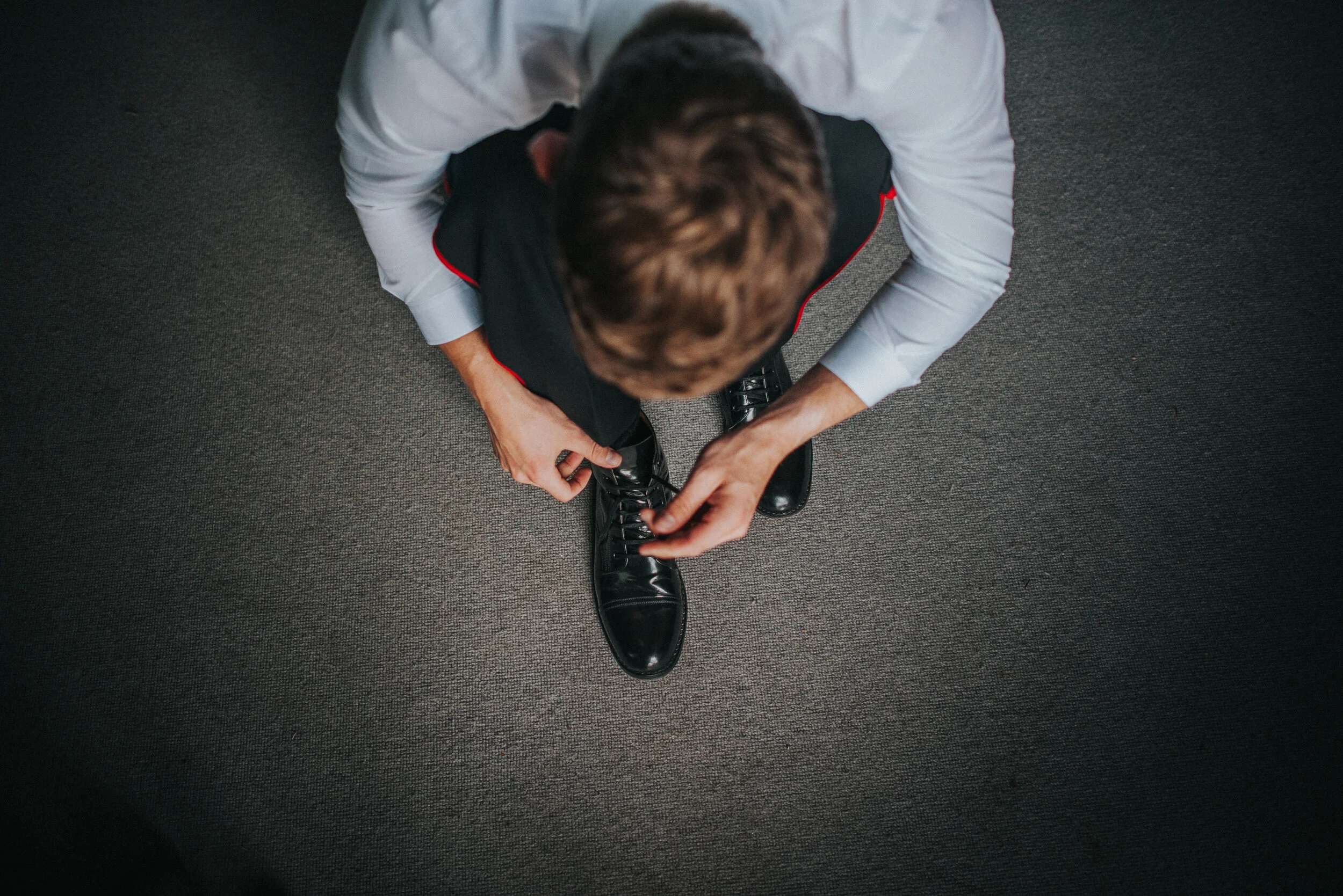 Top-down view of a man in a white shirt tying his black dress shoes while sitting on a dark carpeted floor.
