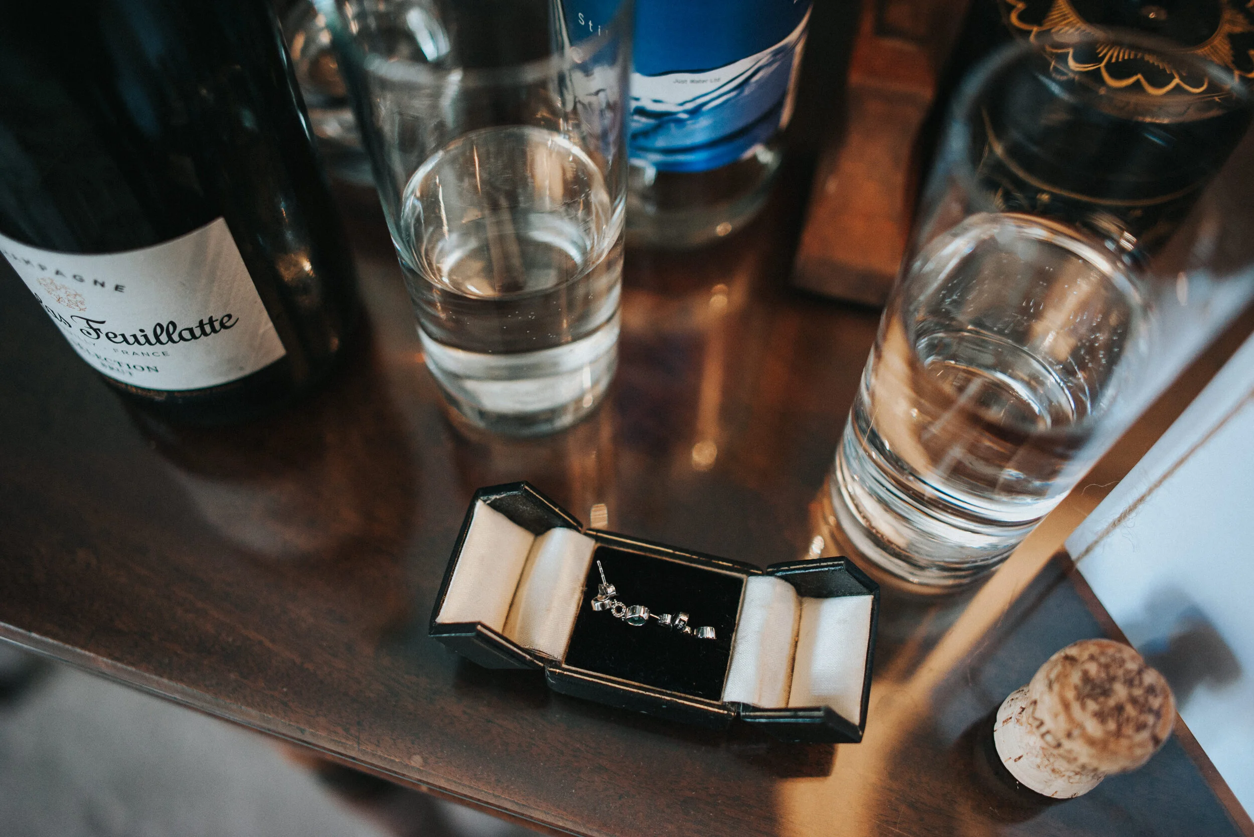 A bottle of red wine, two glasses of water, a small box with a silver necklace that spells out 'Love', a blue bottle, and a cork on a wooden table.