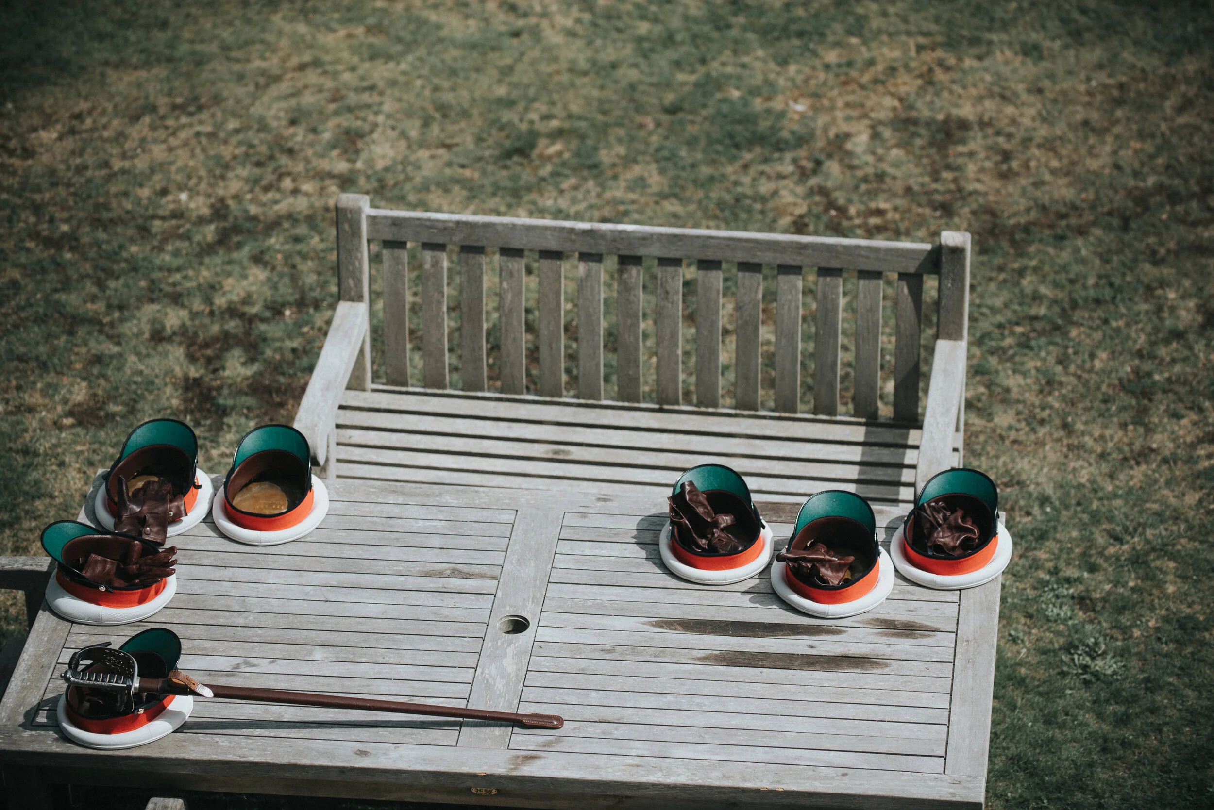 Several pairs of curling shoes and a curling brush placed on a wooden outdoor table, with an empty wooden bench and a grassy area in the background.