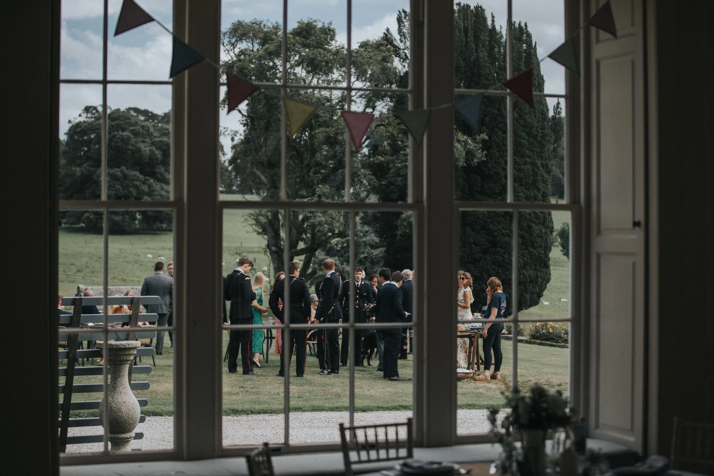 Guests gathering outdoors through a large window decorated with small pennant banners, in a lush green garden setting.