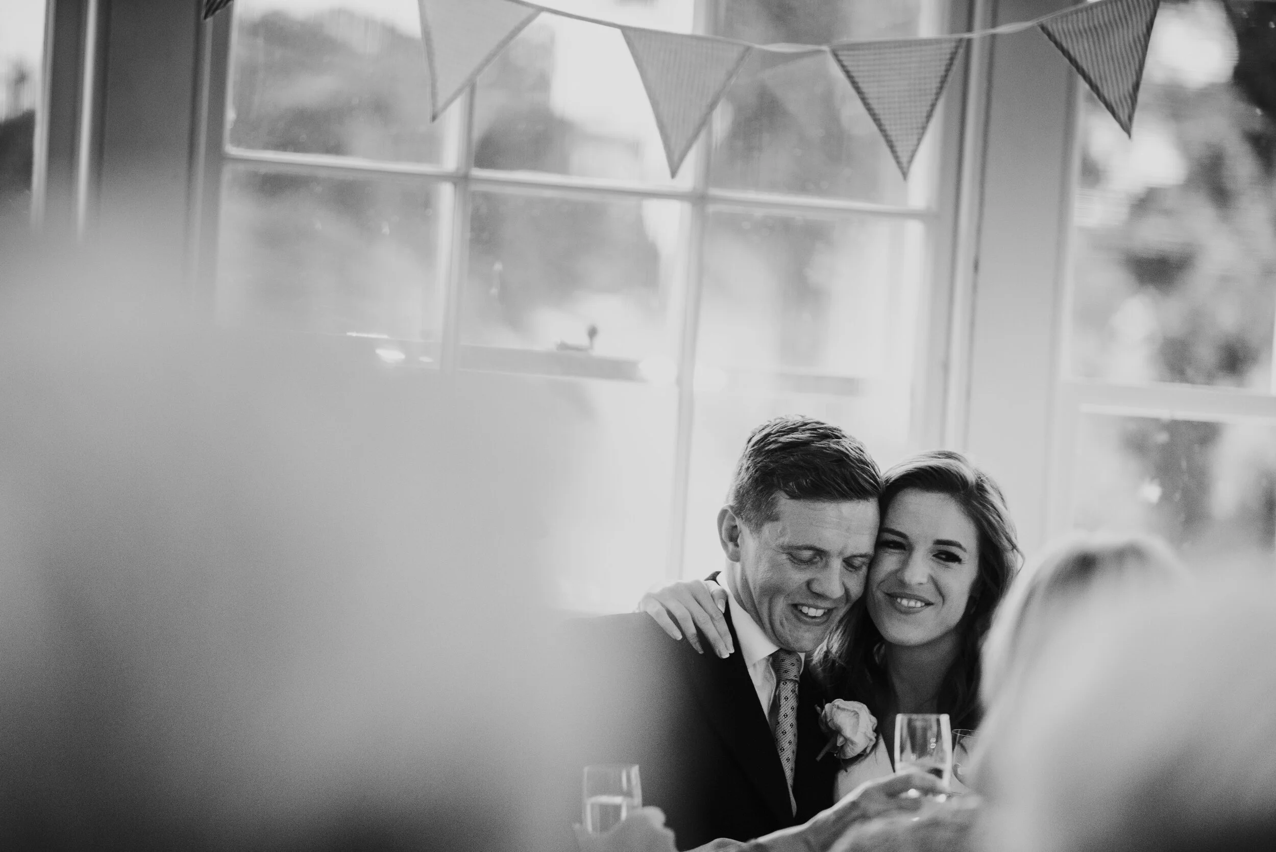Black and white photo of a smiling couple at a celebration, with the man wearing a suit and tie, and the woman with her arm around him, both holding glasses of champagne, sitting in front of a window with bunting decorations.