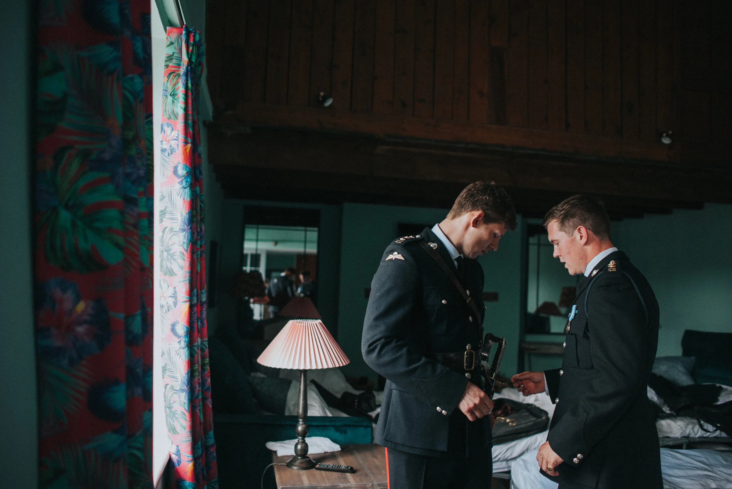 Two military officers standing indoors, looking at an object in one of their hands, in a dimly lit room with a bed, a lamp, and a colorful curtain.