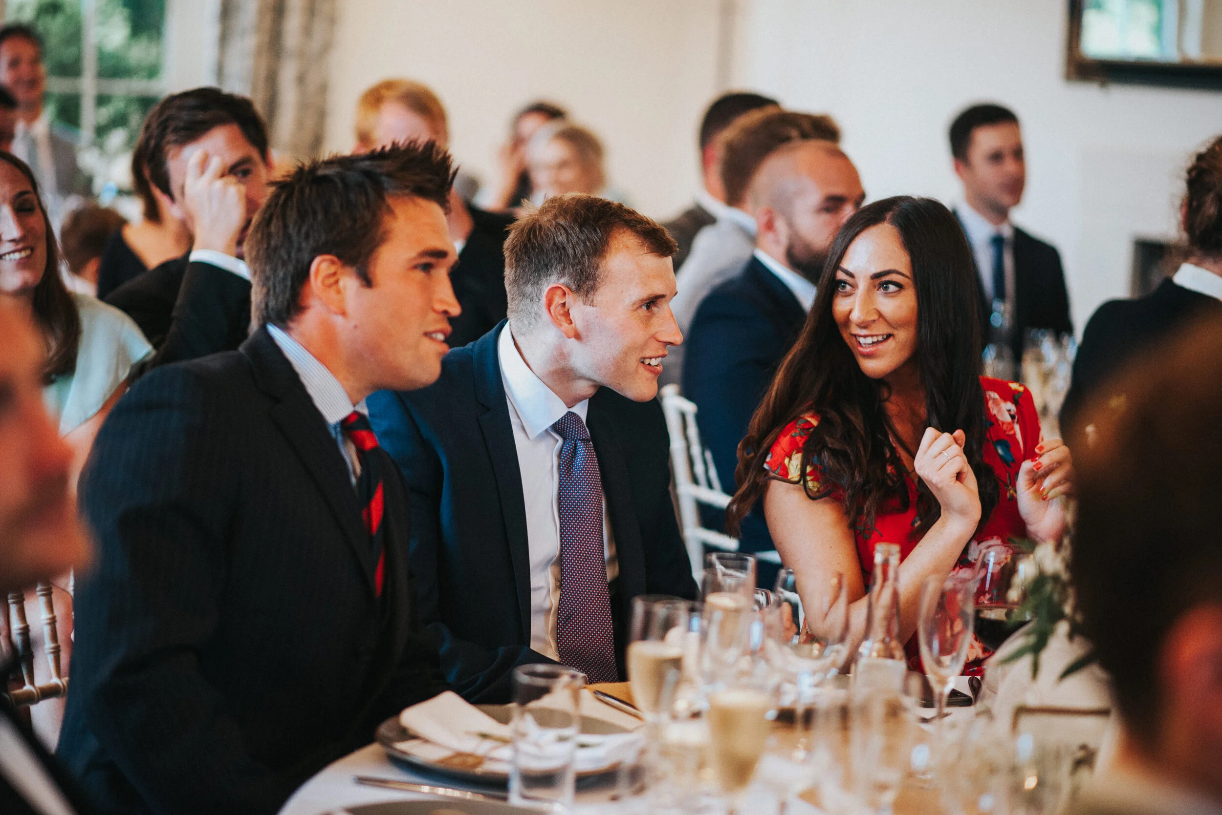 People are sitting at a formal event, dressed in suits and dresses, engaging in conversation and smiling, with a decorated table of drinks and tableware.