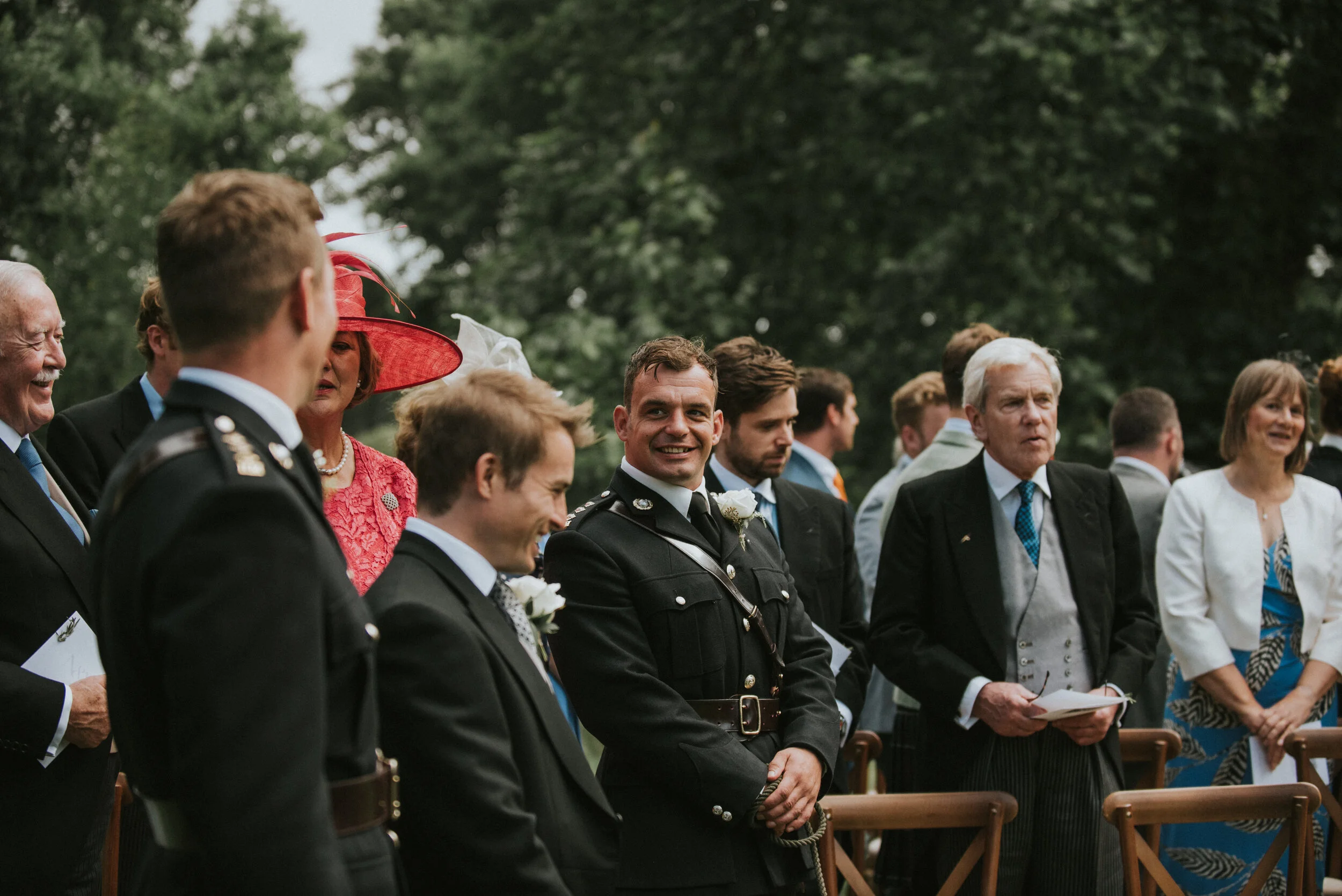 People attending an outdoor wedding ceremony, including men in formal suits and military uniforms, and women in dressy attire, standing among empty wooden chairs with a lush green background.