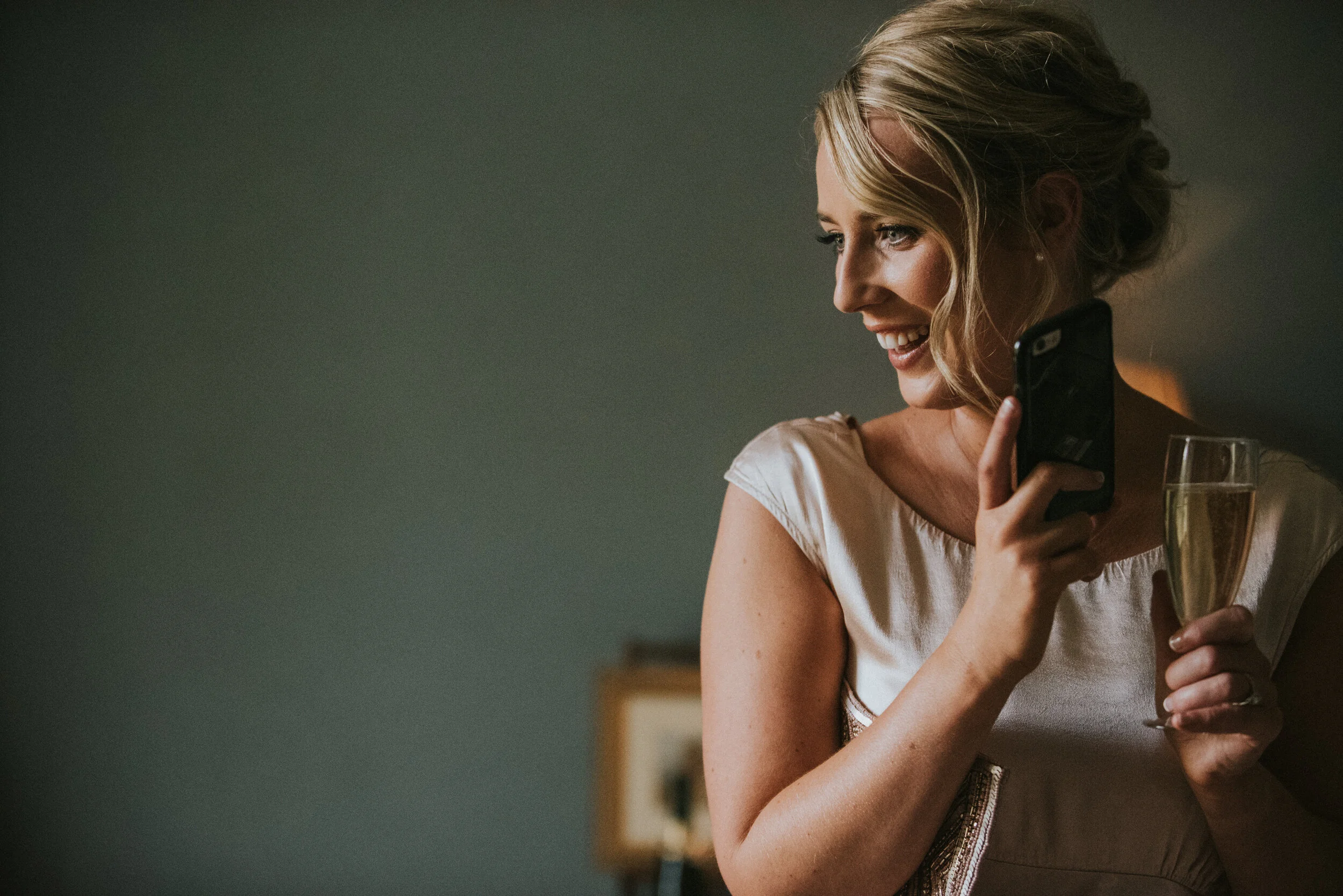 A smiling woman with blonde hair in an updo, holding a glass of champagne and a phone, looking to her right at a celebration.