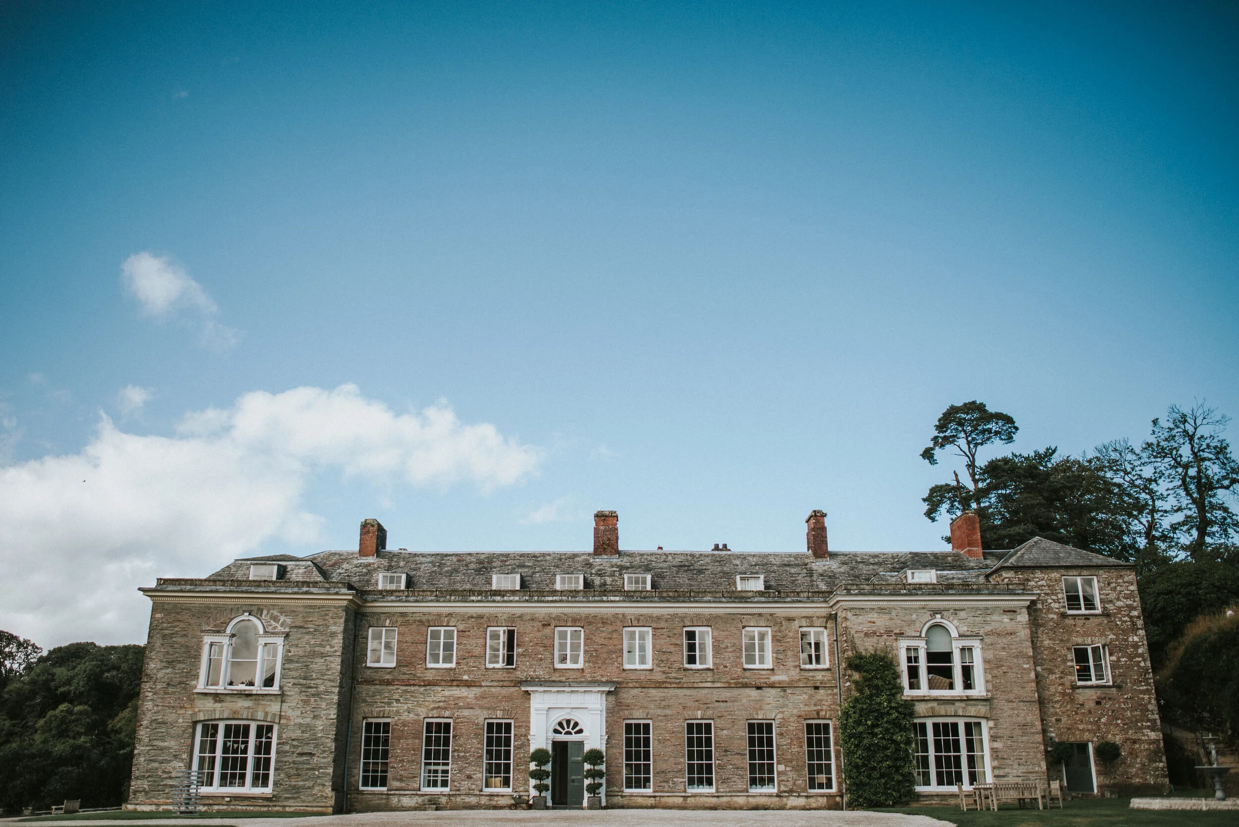 A large stone mansion with multiple windows, doors, and chimneys, surrounded by trees and sky.