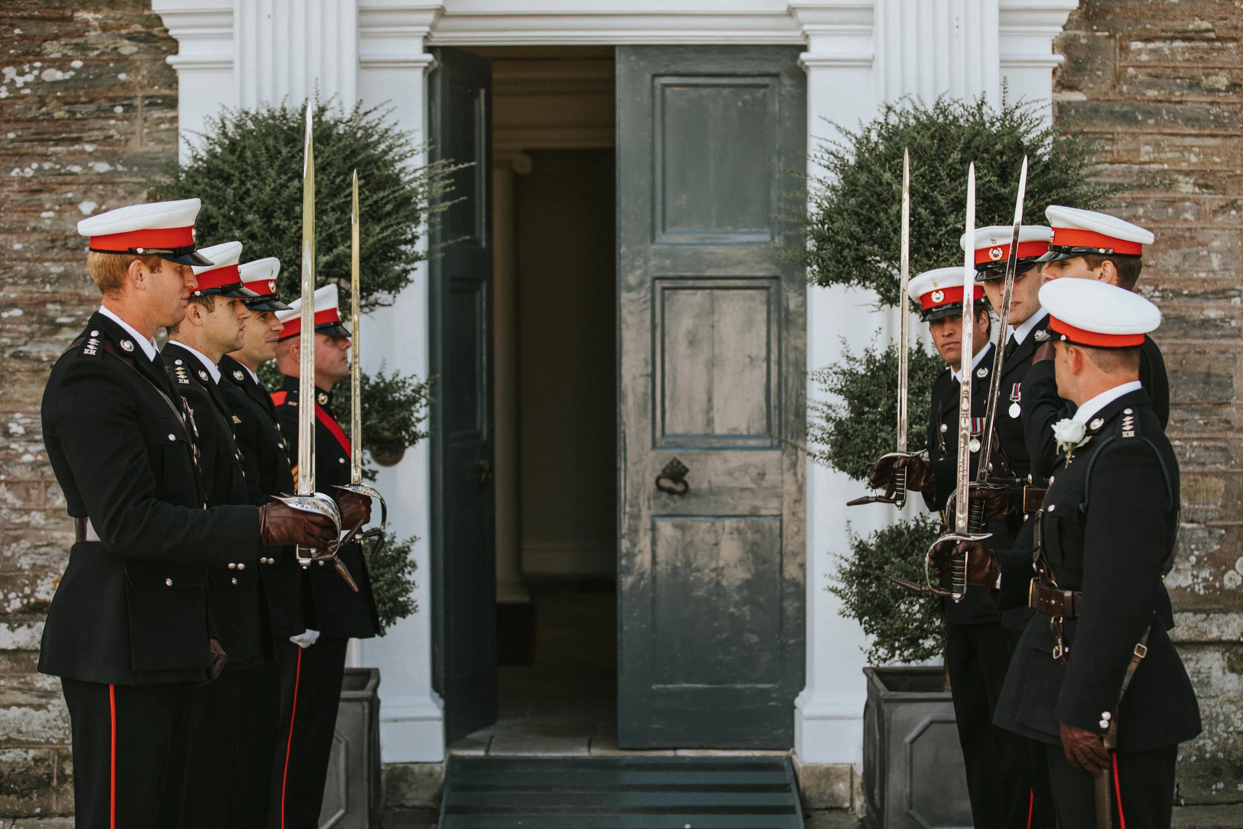 Military honor guard standing in formation outside a building with an open door, with two potted plants on each side.