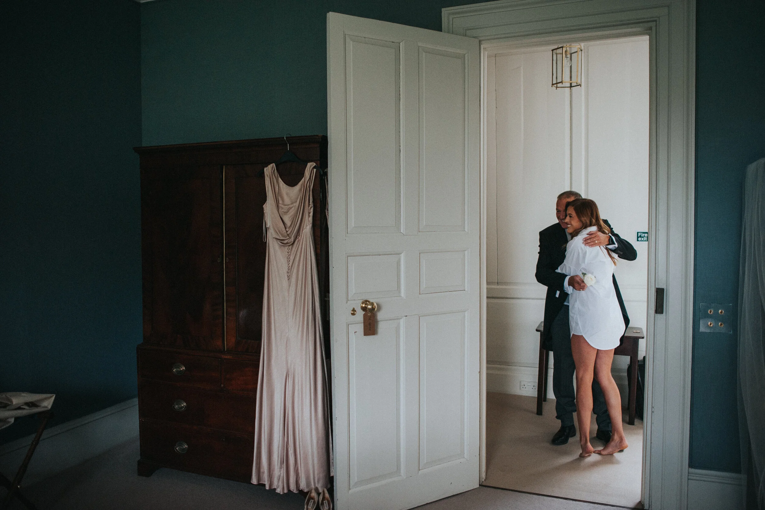 A bride and groom hugging in a room, with the bride wearing a white shirt and the groom in a suit, as seen through an open door.