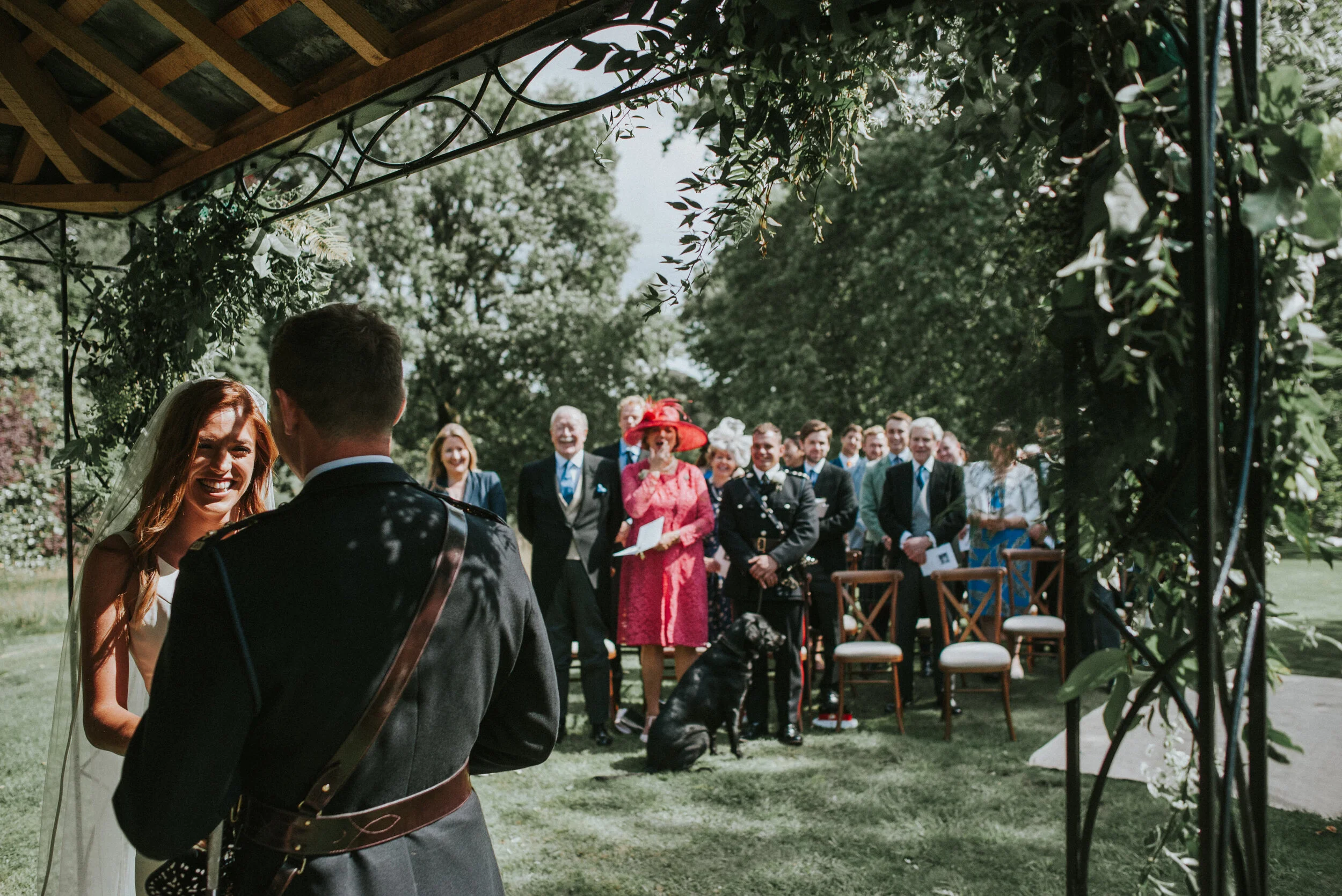 A wedding ceremony outdoors with a bride and groom standing under a decorated arch. The bride is smiling and facing the groom, who has his back to the camera. Guests are seated and standing in the background, including a woman in a bright pink dress 