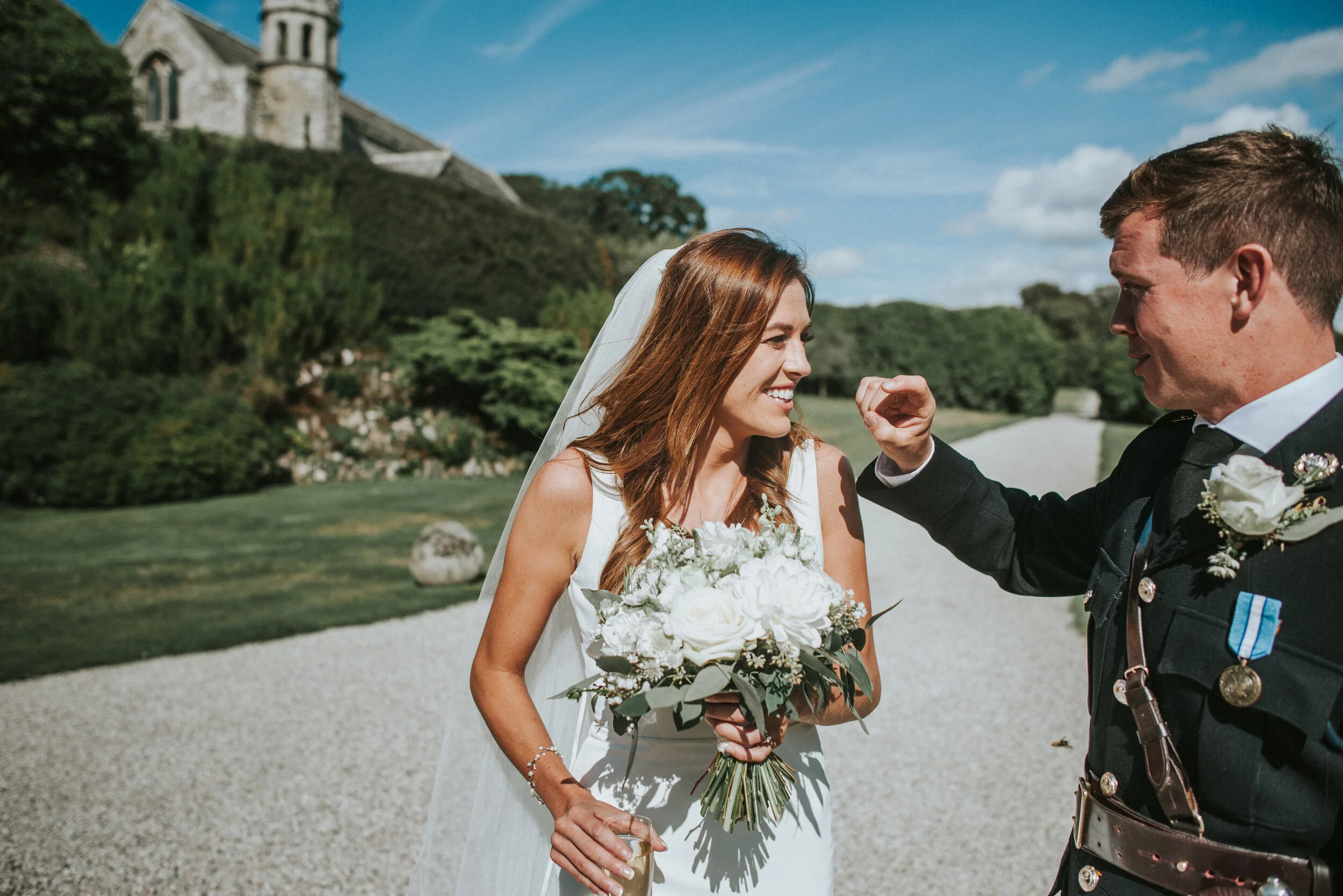A bride and groom sharing a moment outdoors on their wedding day, with the bride wearing a white dress and holding a bouquet, and the groom in a black uniform, in front of a scenic landscape with a church and greenery.