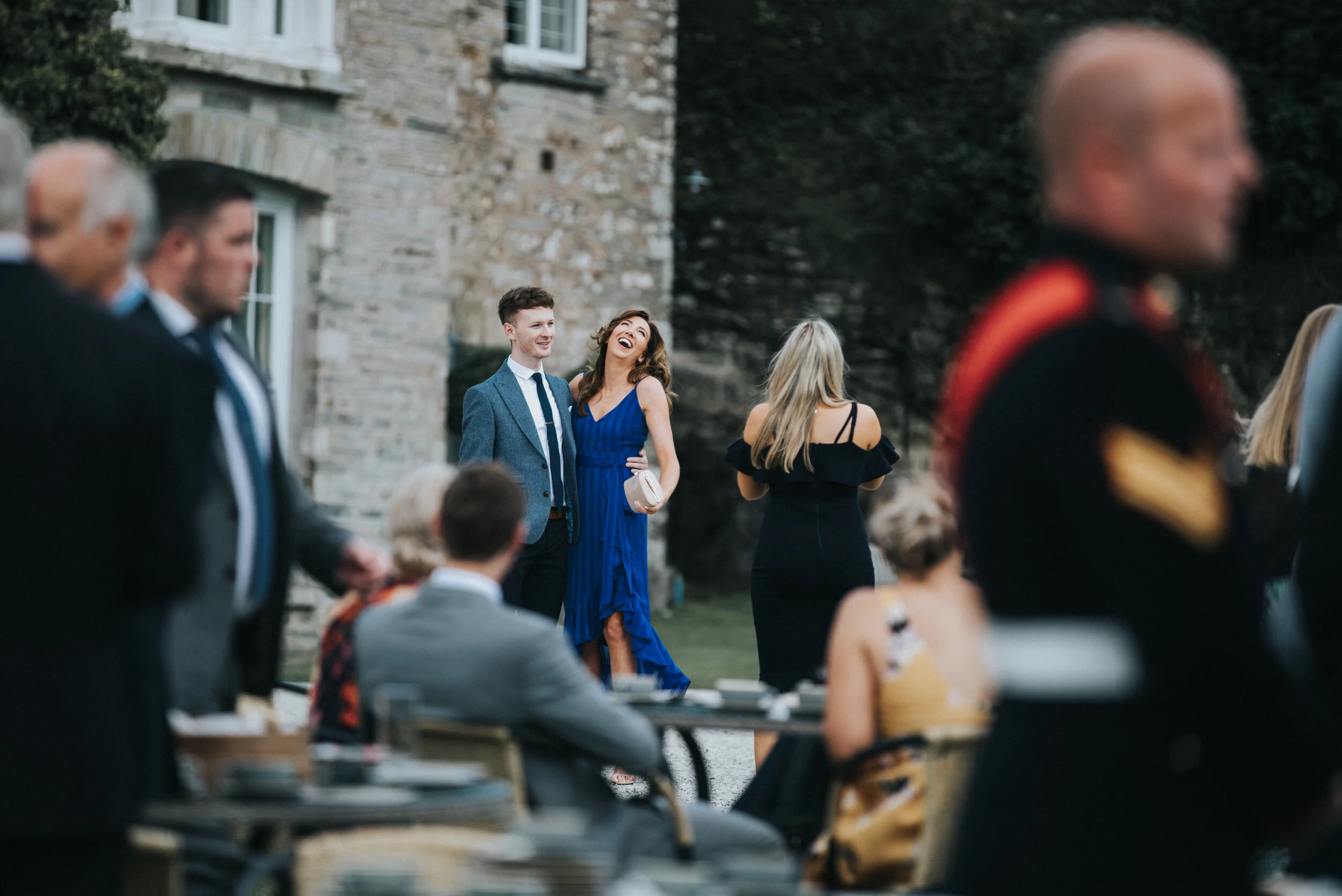 People socializing at an outdoor event near a stone building, with a woman in a blue dress laughing and a man in a gray suit standing nearby.