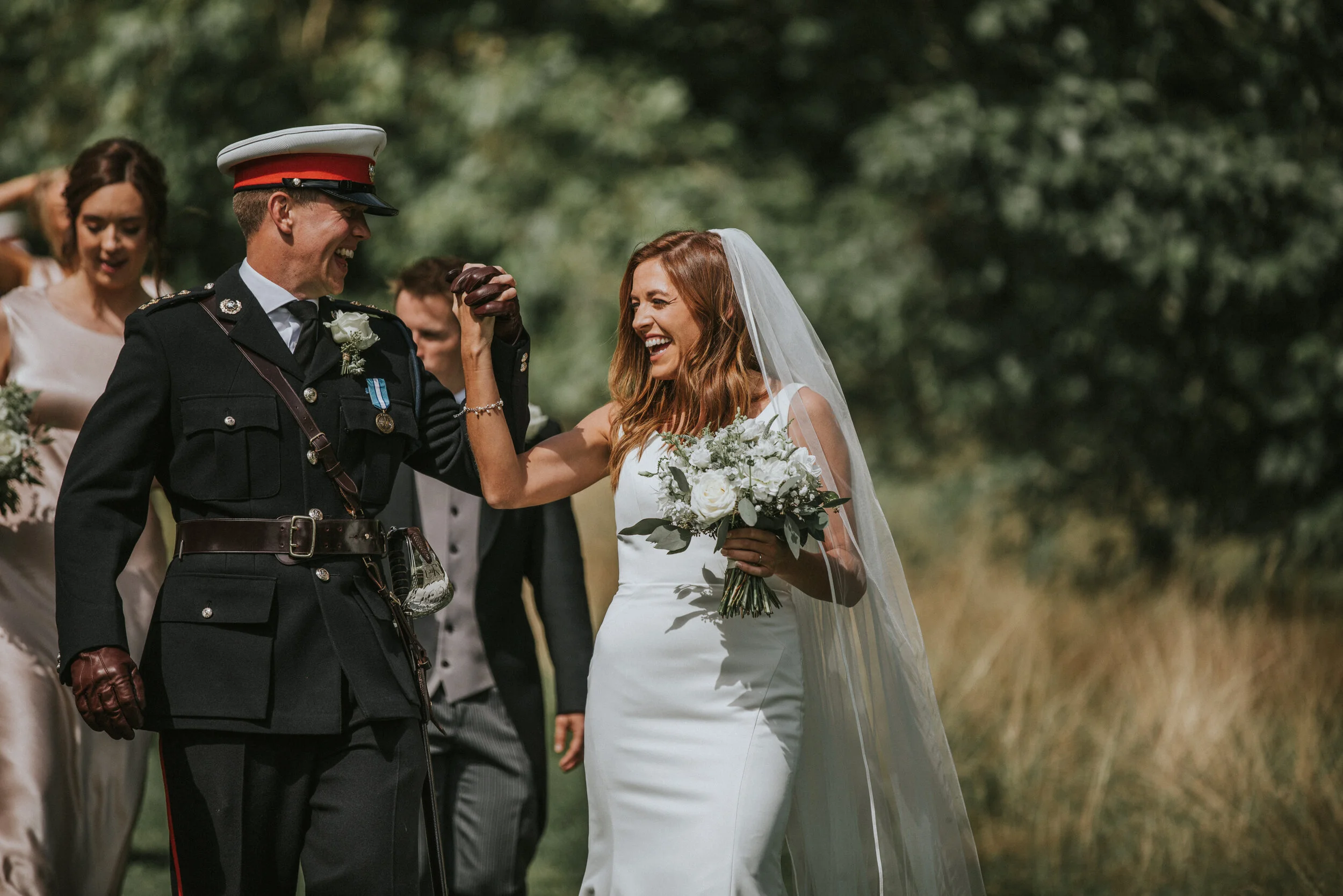 A bride and a uniformed military officer are celebrating outdoors, smiling and fist-bumping, with other wedding guests in the background.