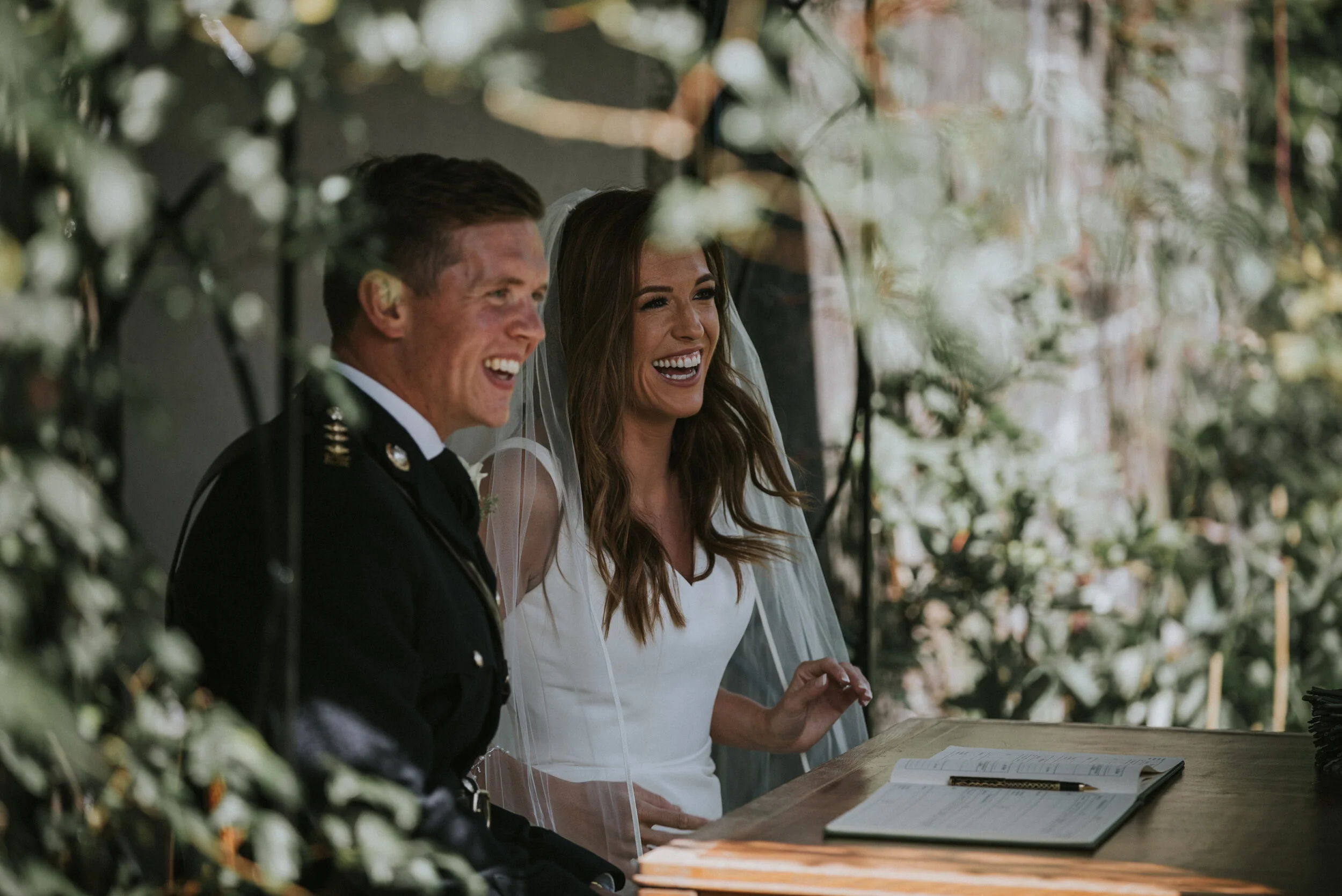 A bride and groom seated at a table, smiling, during a wedding ceremony in a decorated outdoor setting surrounded by green foliage.