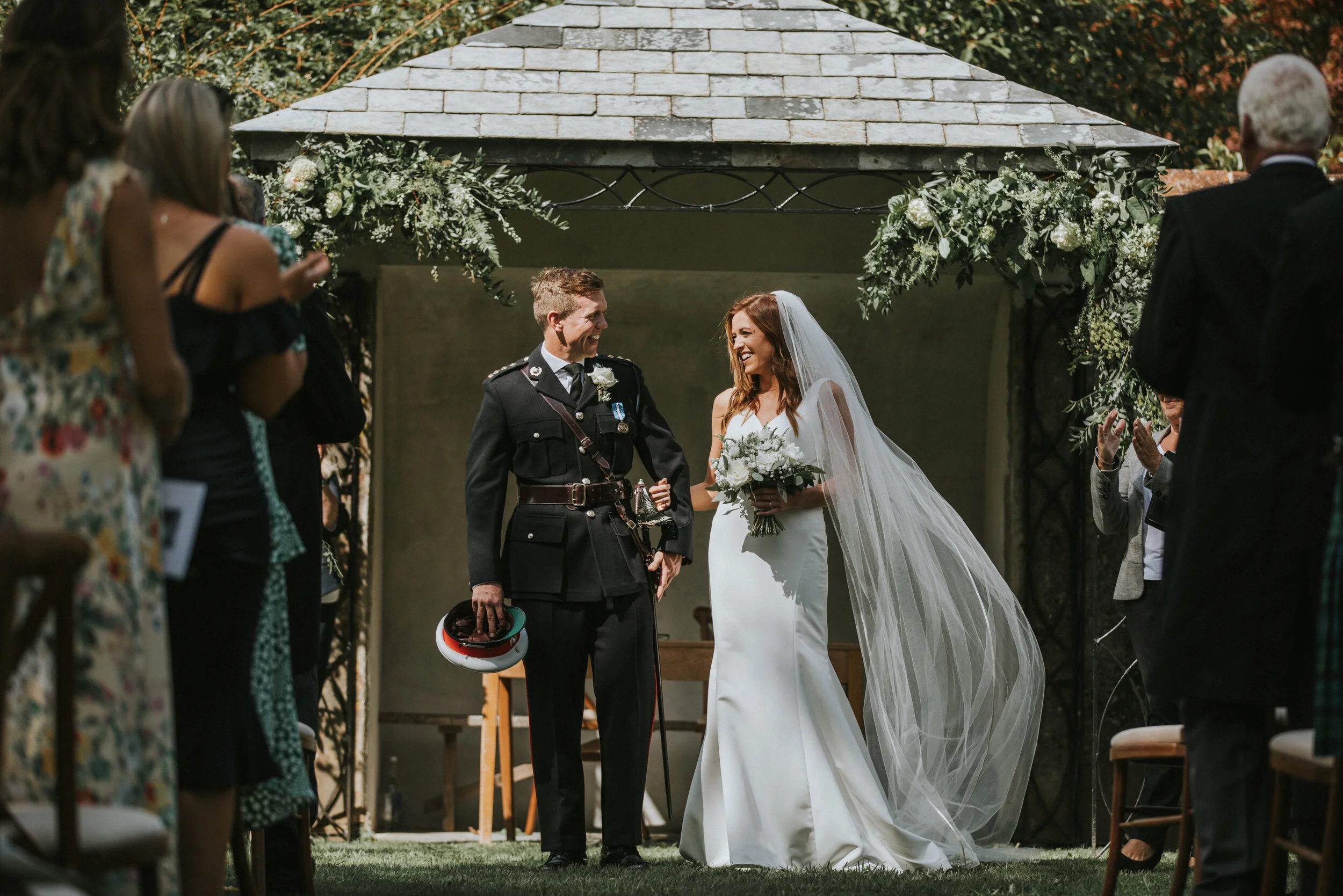 A bride in a white gown and veil stands with a groom in a military uniform during a wedding ceremony outdoors. They are smiling and holding hands, surrounded by guests clapping and celebrating under a decorated pavilion.