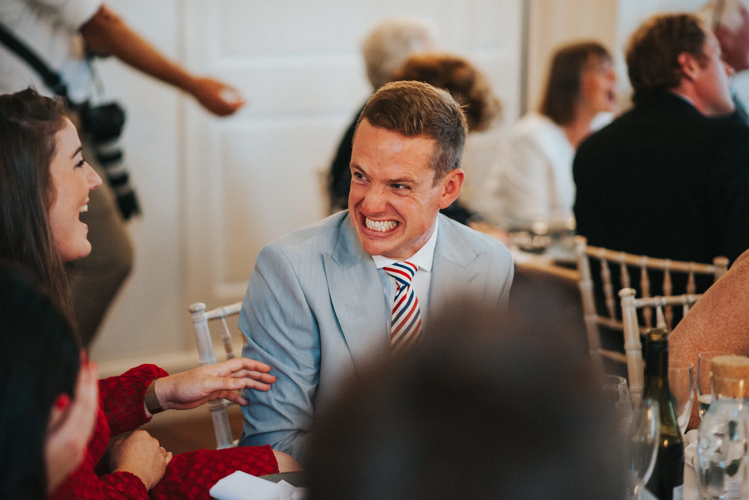 People sitting around a table at a social gathering, smiling and engaging in conversation, with a man in a light gray suit and a striped tie smiling broadly.