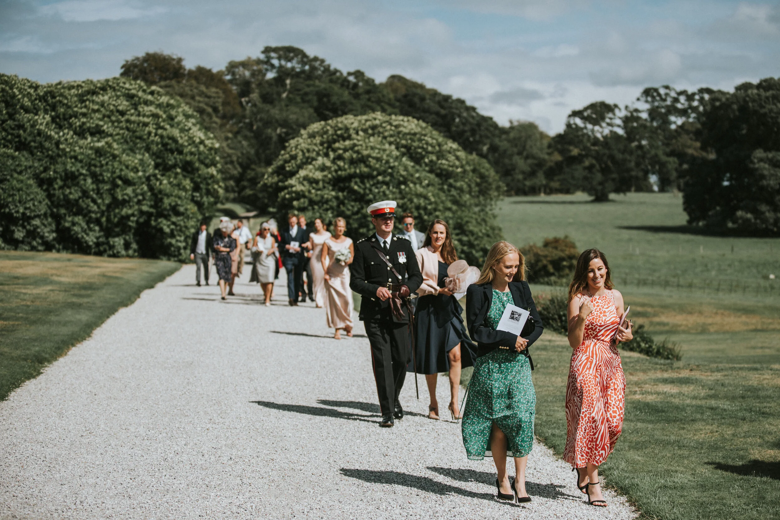 A group of people, including a man in a formal military uniform, walking on a gravel path in a park-like setting with trees and grass, during a sunny day.