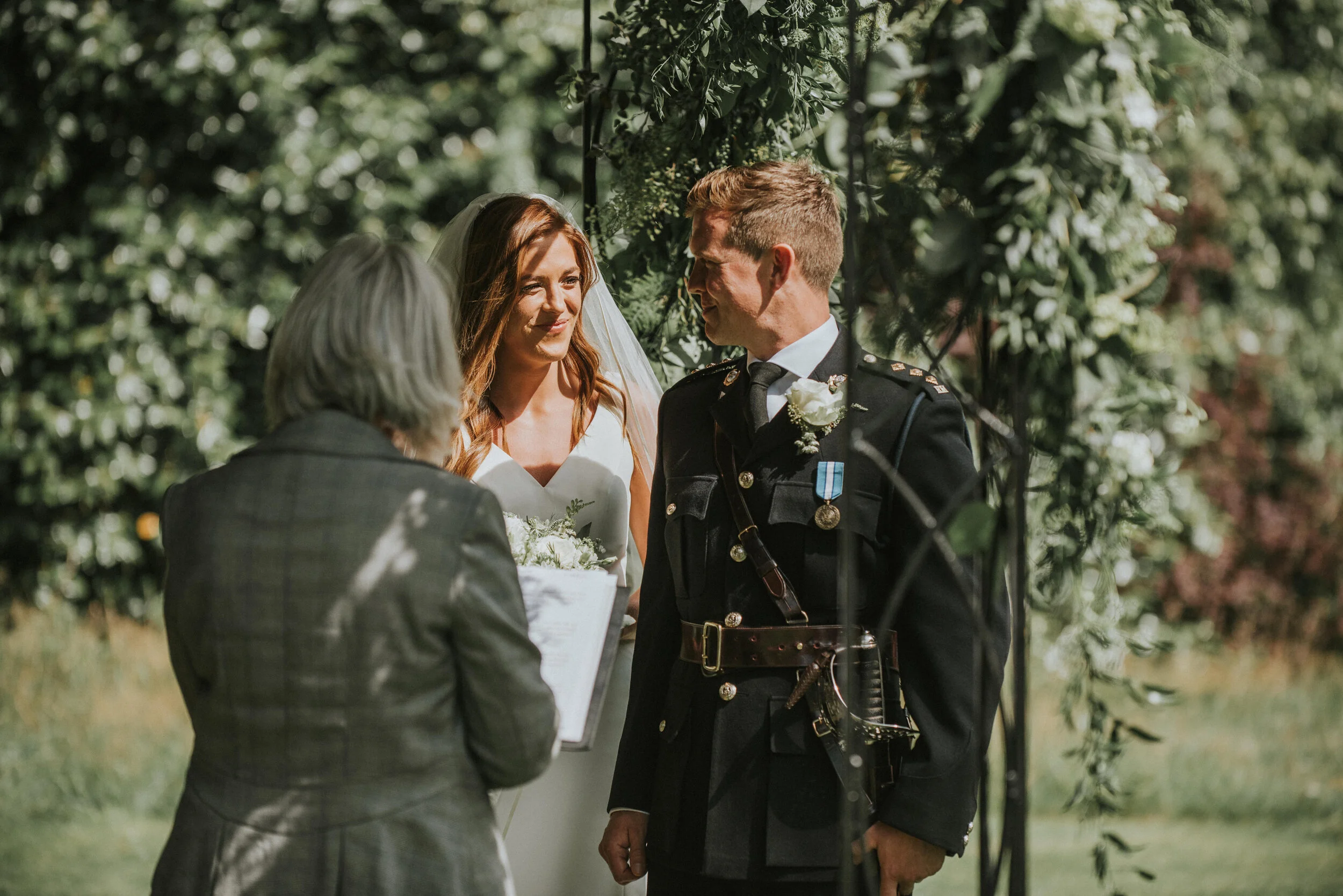 A wedding ceremony taking place outdoors with a bride, groom in a military uniform, and officiant during the vows.