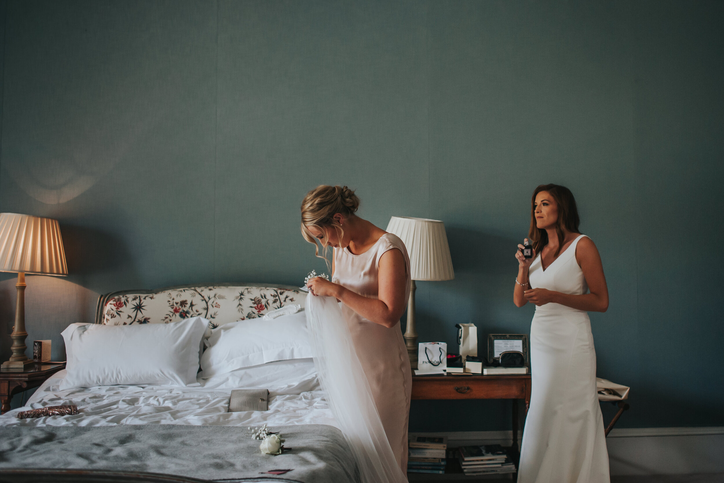 Two women in white dresses preparing in a bedroom, with one holding a wedding dress and the other holding a perfume bottle.