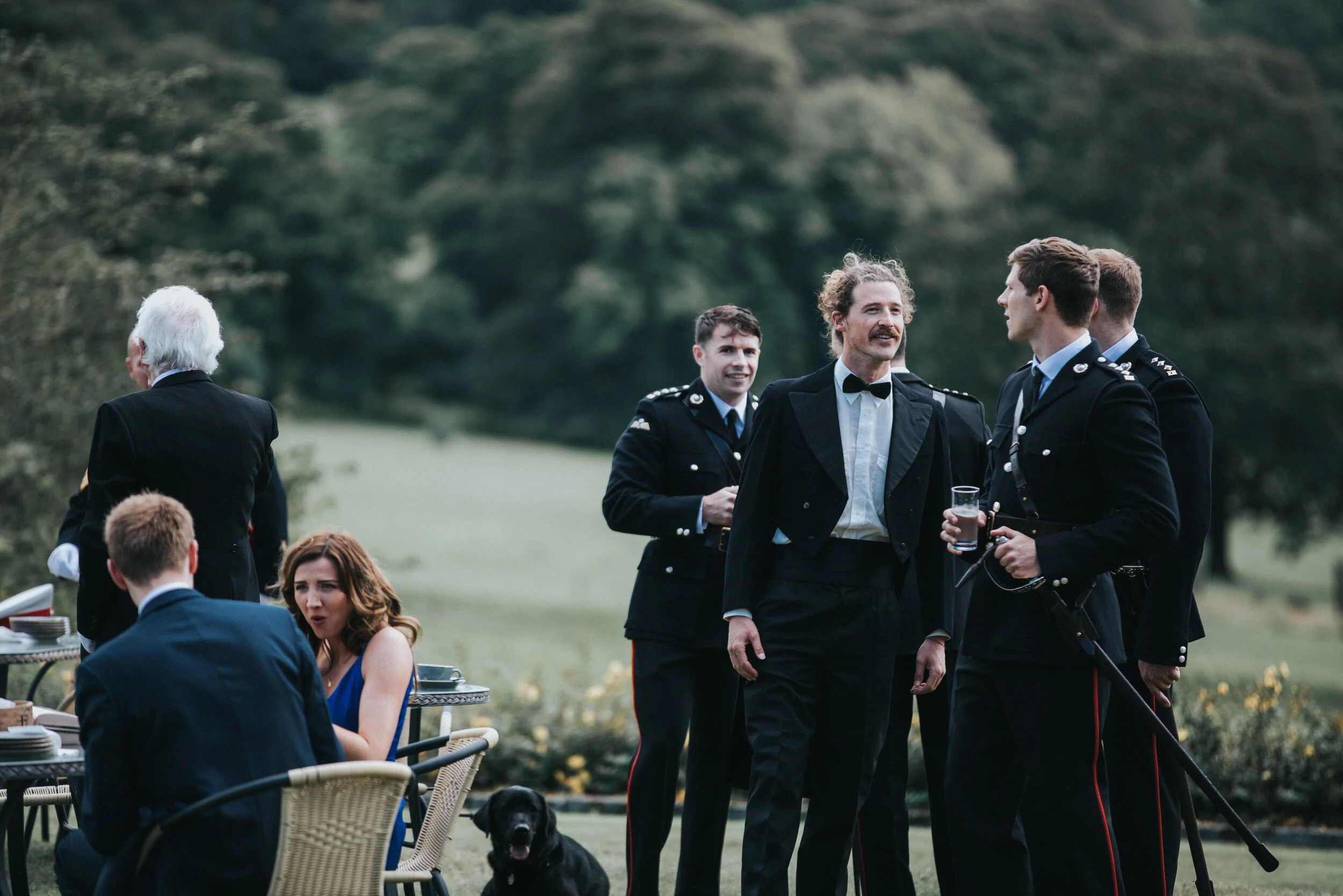 People dressed in formal attire, including military uniforms, socializing outdoors in a grassy area with trees in the background, with a woman sitting at a table and a black dog nearby.