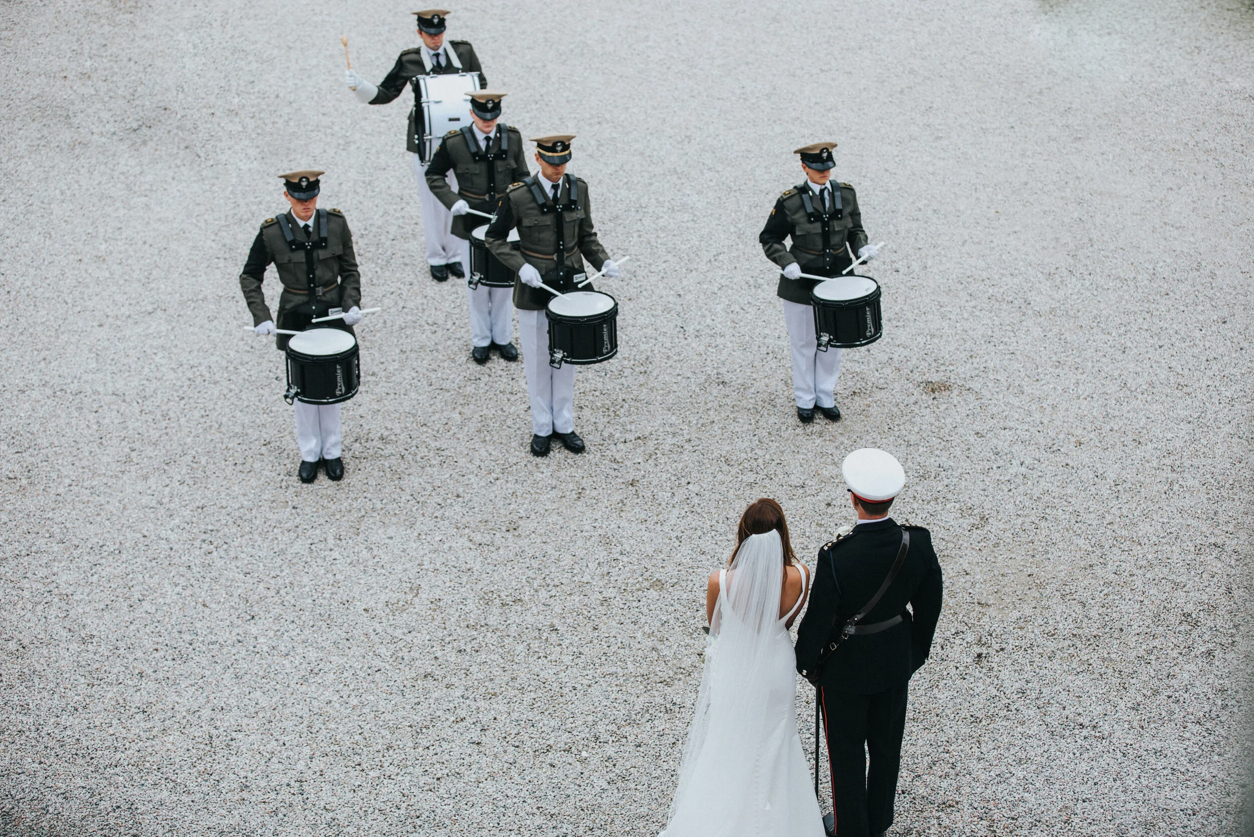 A bride and groom standing outdoors, facing a military marching band performing a ceremony, with the band members playing drums and wearing military uniforms.