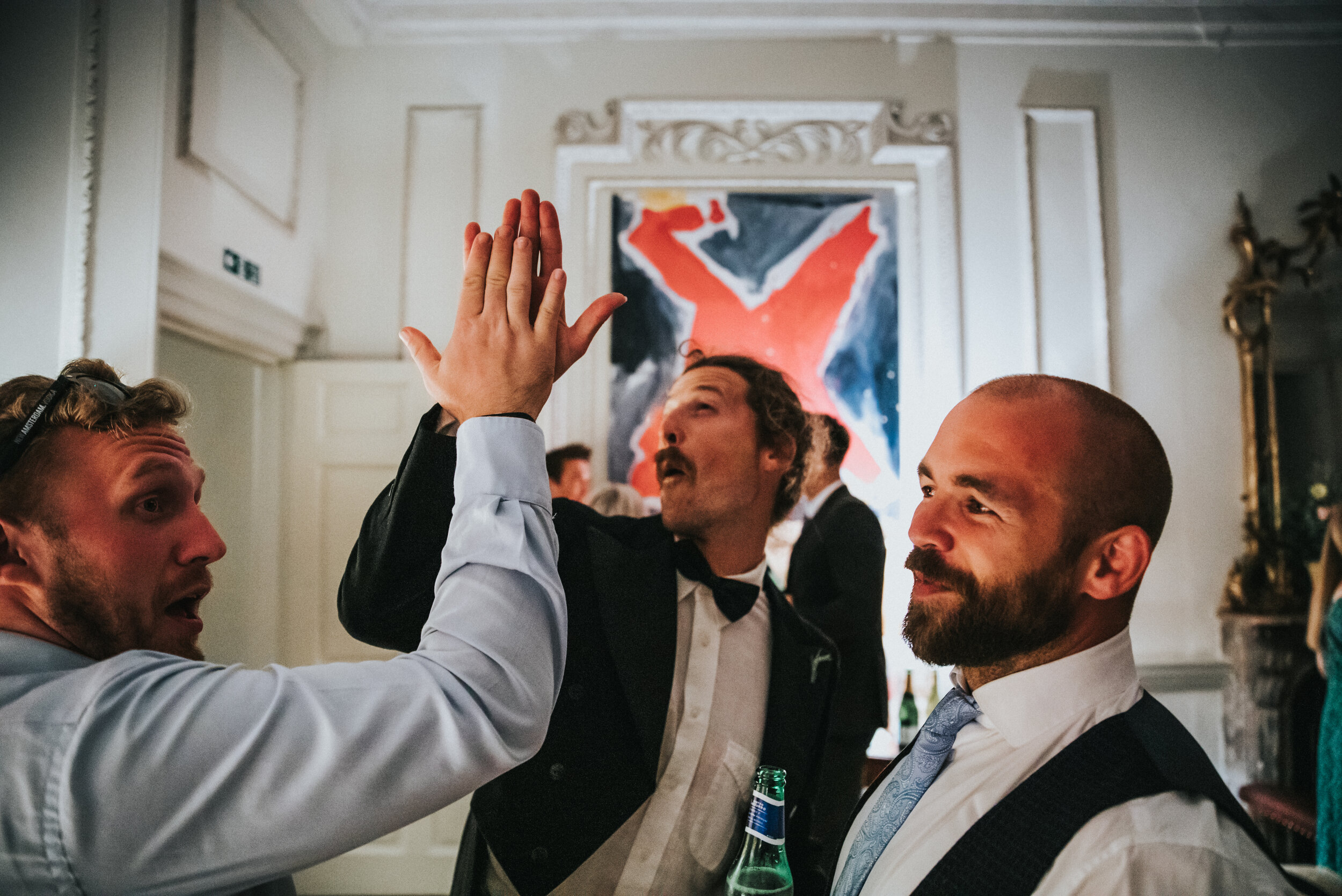 Three men in formal attire exchanging a high five at a party in an elegant room with artwork on the wall.