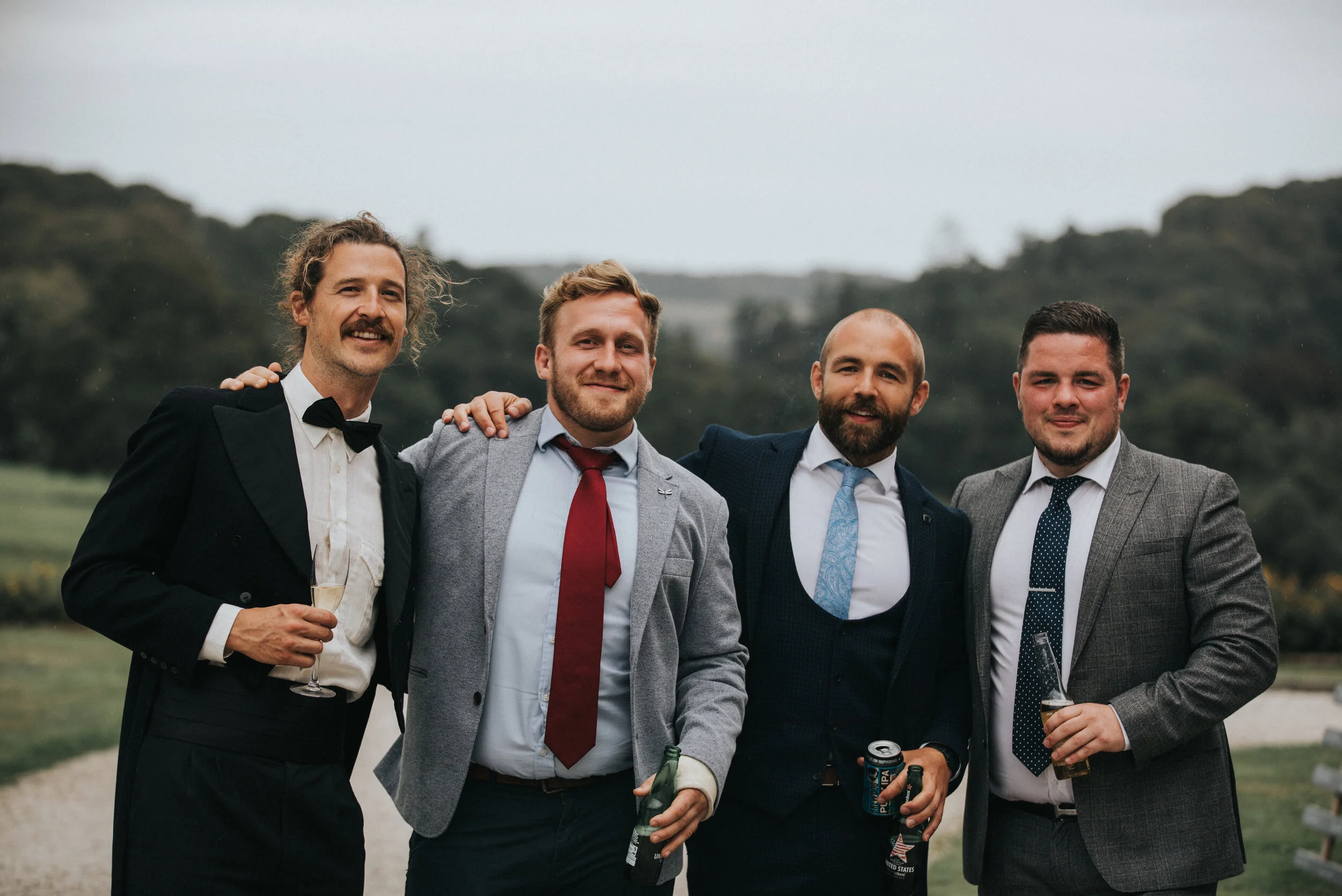 Four men dressed in formal attire standing outdoors with arms around each other, smiling, holding drinks in their hands, on a cloudy day with greenery in the background.