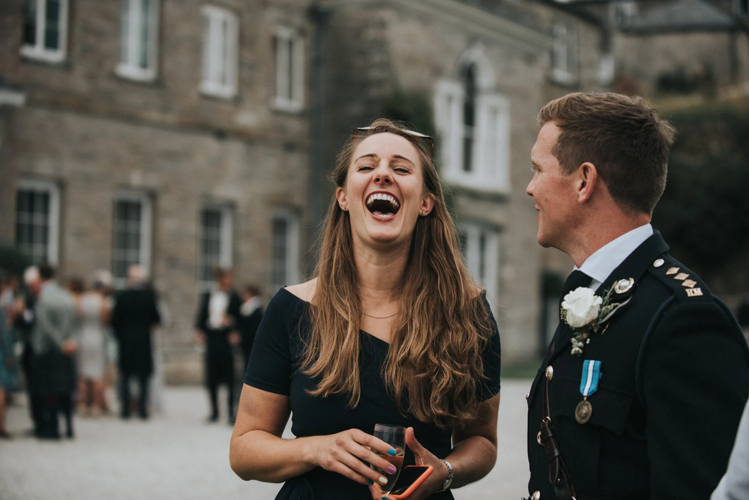 A woman with long, wavy brown hair laughing and holding a glass, standing next to a man in a military uniform at an outdoor event with a stone building and other guests in the background.