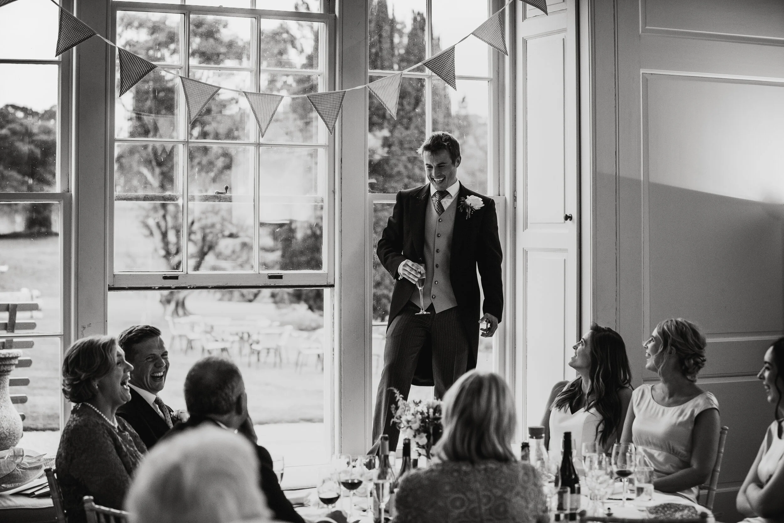 A group of people at a wedding reception, with a man in a suit standing in front of a large window, holding a glass and smiling, while others seated at a table look and laugh.