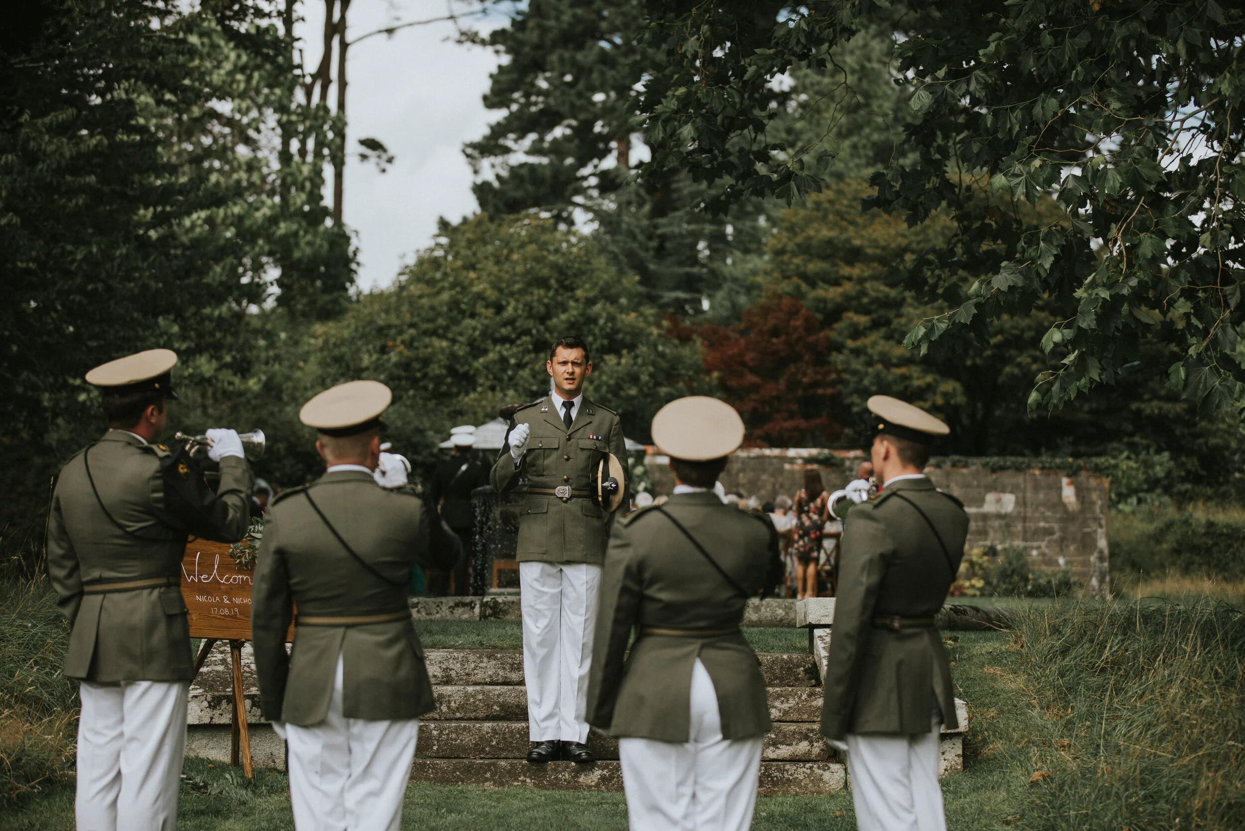 A military funeral ceremony outdoors with a military officer giving a speech, and five uniformed soldiers saluting nearby, in a green park with trees.