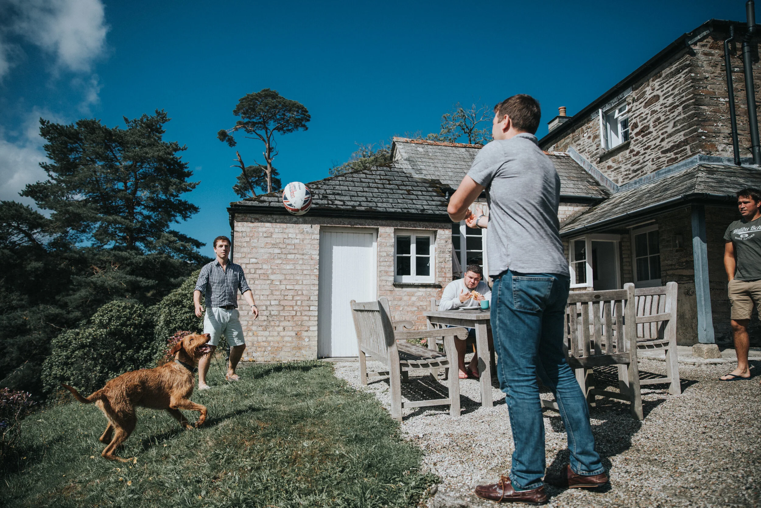 A group of people playing with a soccer ball outside a house on a sunny day. One person is kicking the ball, a dog is running towards it, and other people are sitting and standing near the house, which has a brick exterior and a patio table.