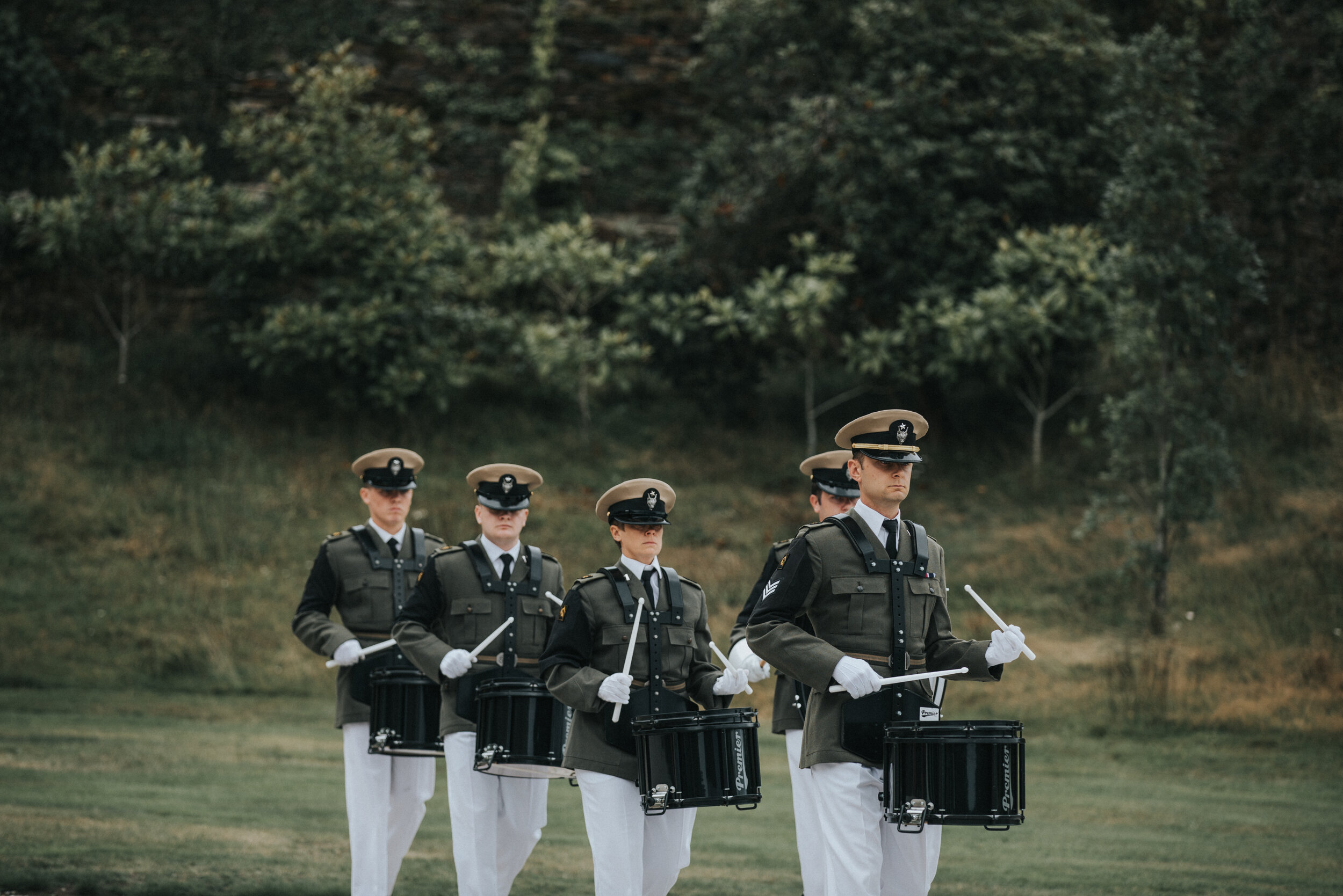 A group of six uniformed marching band members playing drums outdoors on a grassy field with trees in the background.