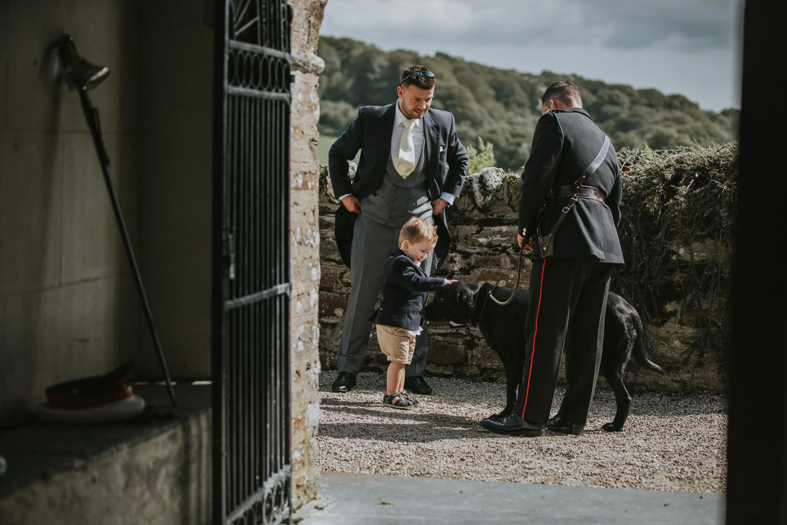 A young boy petting a black service dog for a man in uniform while a man in a suit looks on on a sunny day, with a stone wall and green landscape in the background.