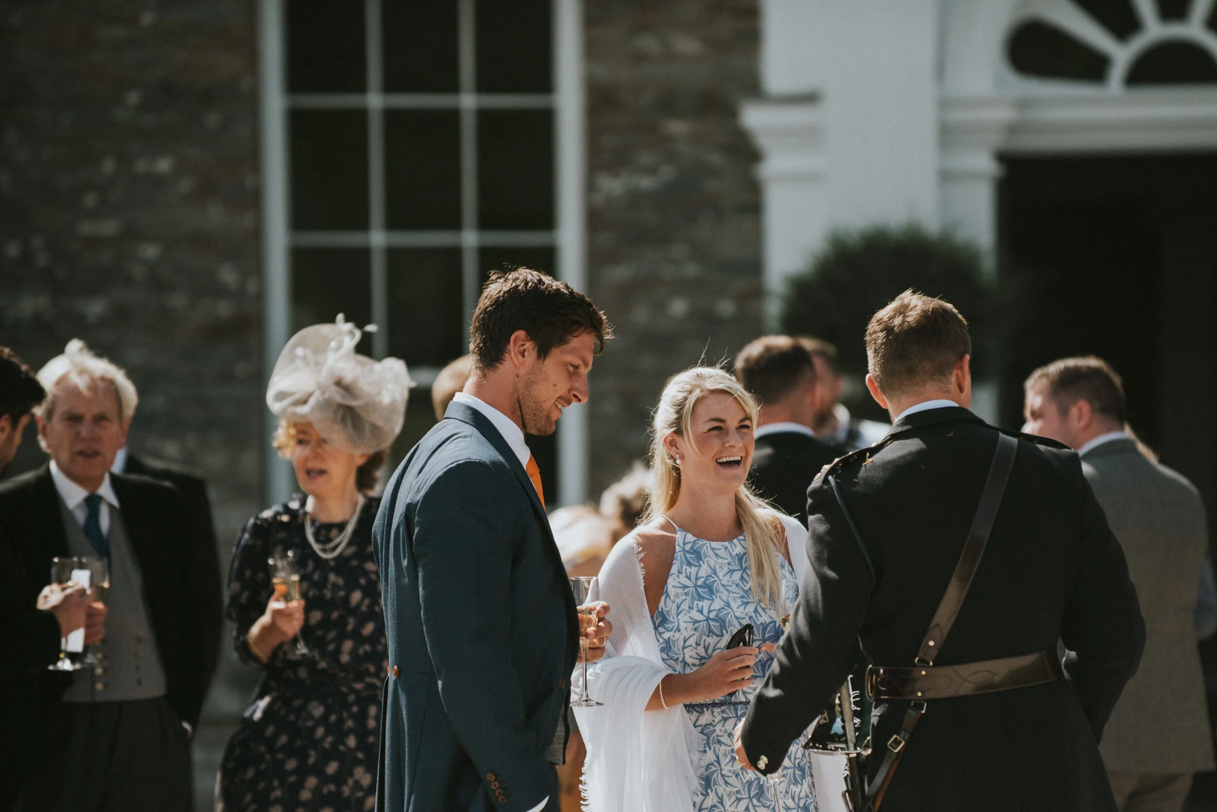 People dressed in formal attire at an outdoor event, smiling and holding drinks, with a building and large window in the background.