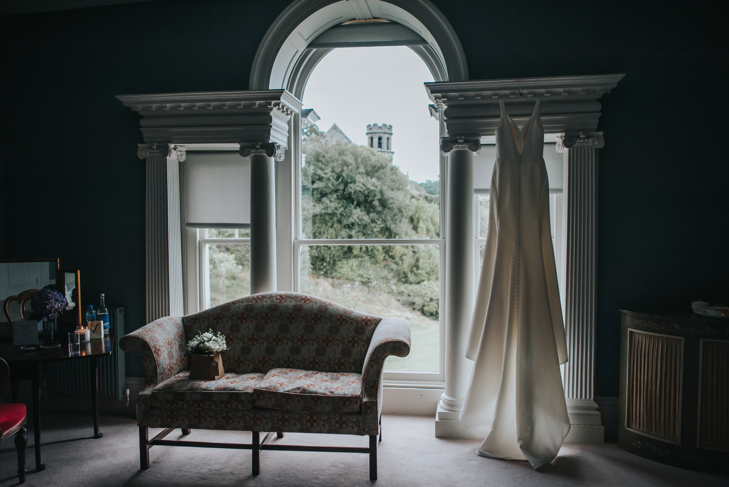 A wedding dress hangs in front of a large window with a view of green trees and a castle tower outside. The room has dark walls, a floral-patterned sofa, a floral vase, and a small table with some bottles and flowers.