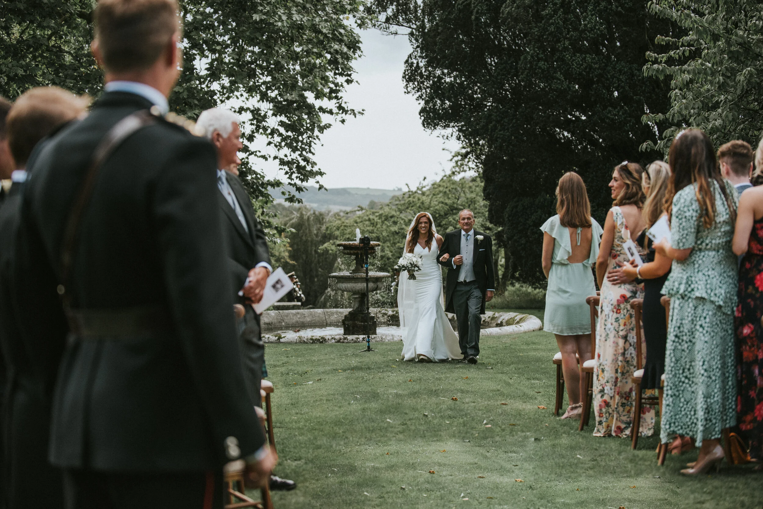 Bride walking down the aisle with her father at an outdoor wedding ceremony, surrounded by seated guests and greenery.