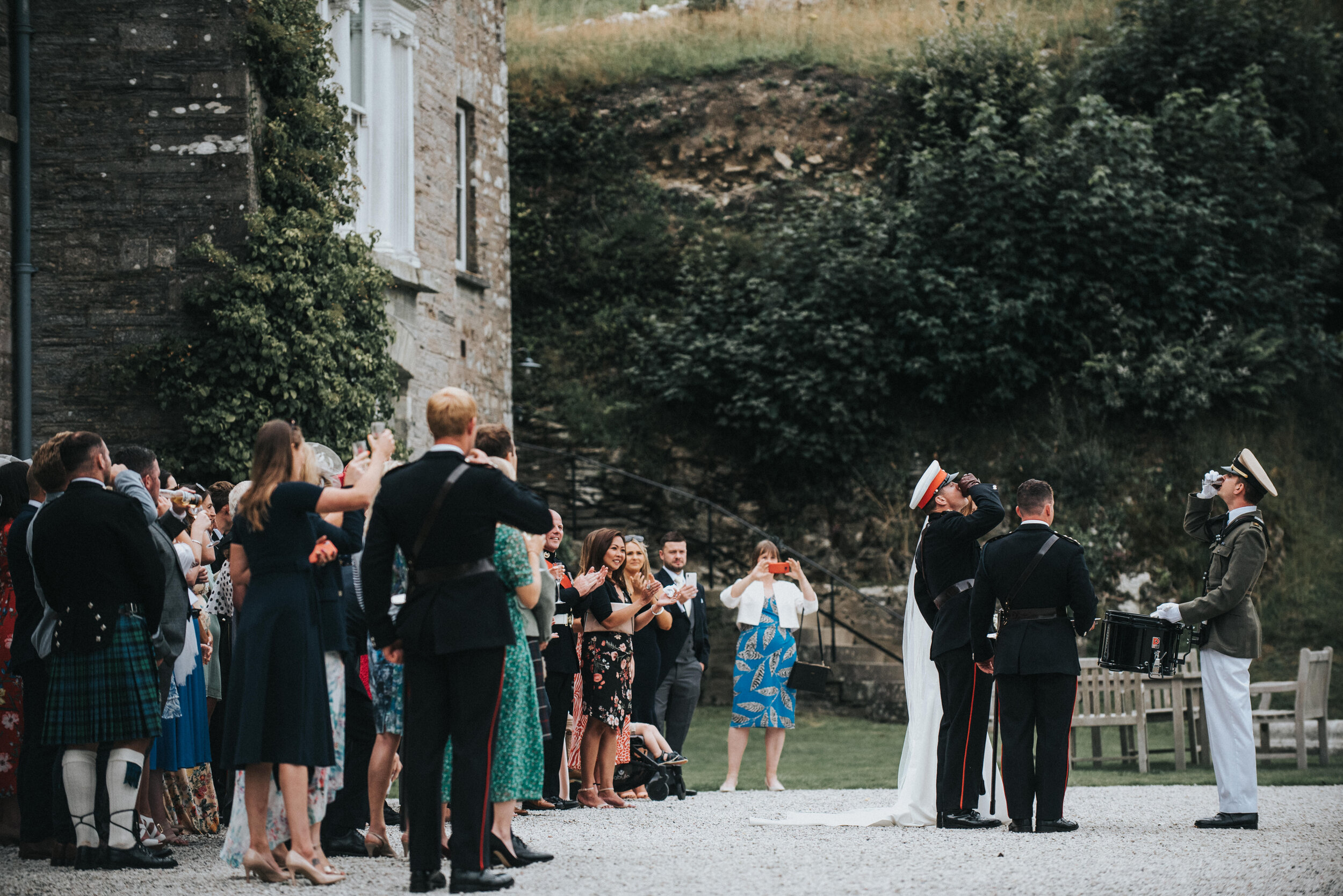 Couple sharing a first dance at a wedding ceremony outdoors, surrounded by guests taking photos and videos, with greenery and a stone building in the background.