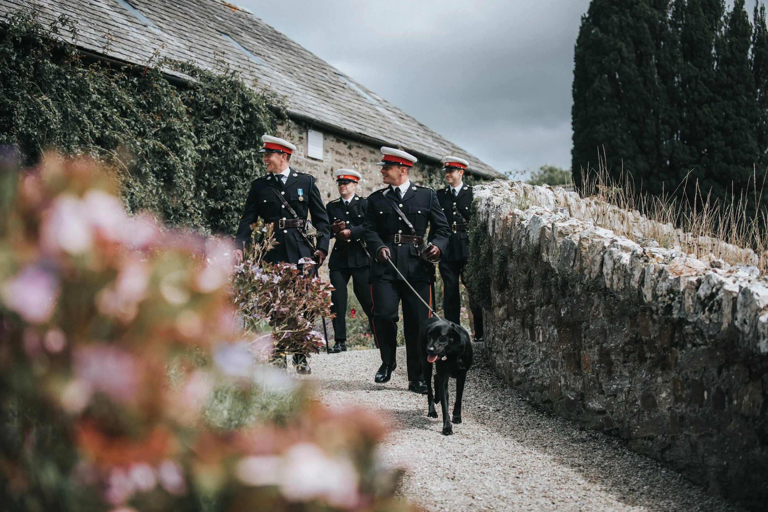 Four police officers in uniform walking outdoors on a gravel path, one policeman with a police dog on a leash, near a stone wall, with a cloudy sky and trees in the background.