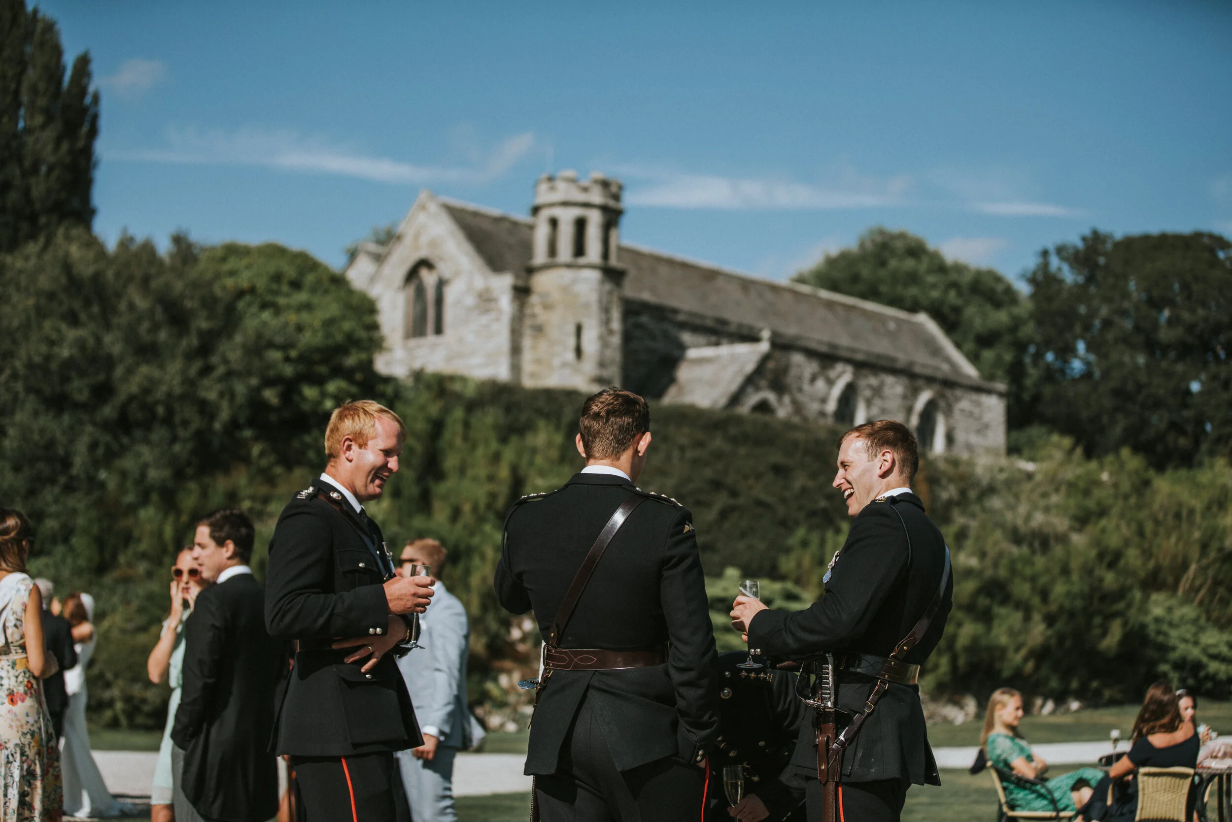 Three men in military uniforms sharing a laugh and holding glasses at an outdoor event, with another group of people and a historic stone building with a tower in the background.