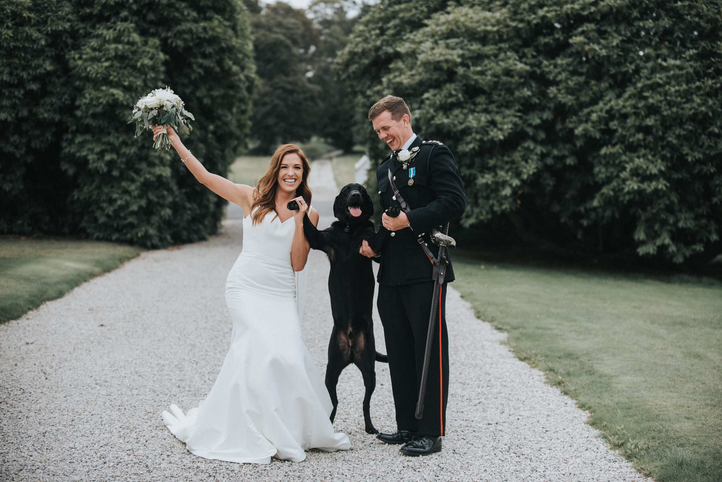 A bride and groom celebrate their wedding with a police dog. The bride wears a white wedding dress and holds a bouquet. The groom is dressed in a military or police uniform. They are both happily smiling, with the dog standing on its hind legs, betwe