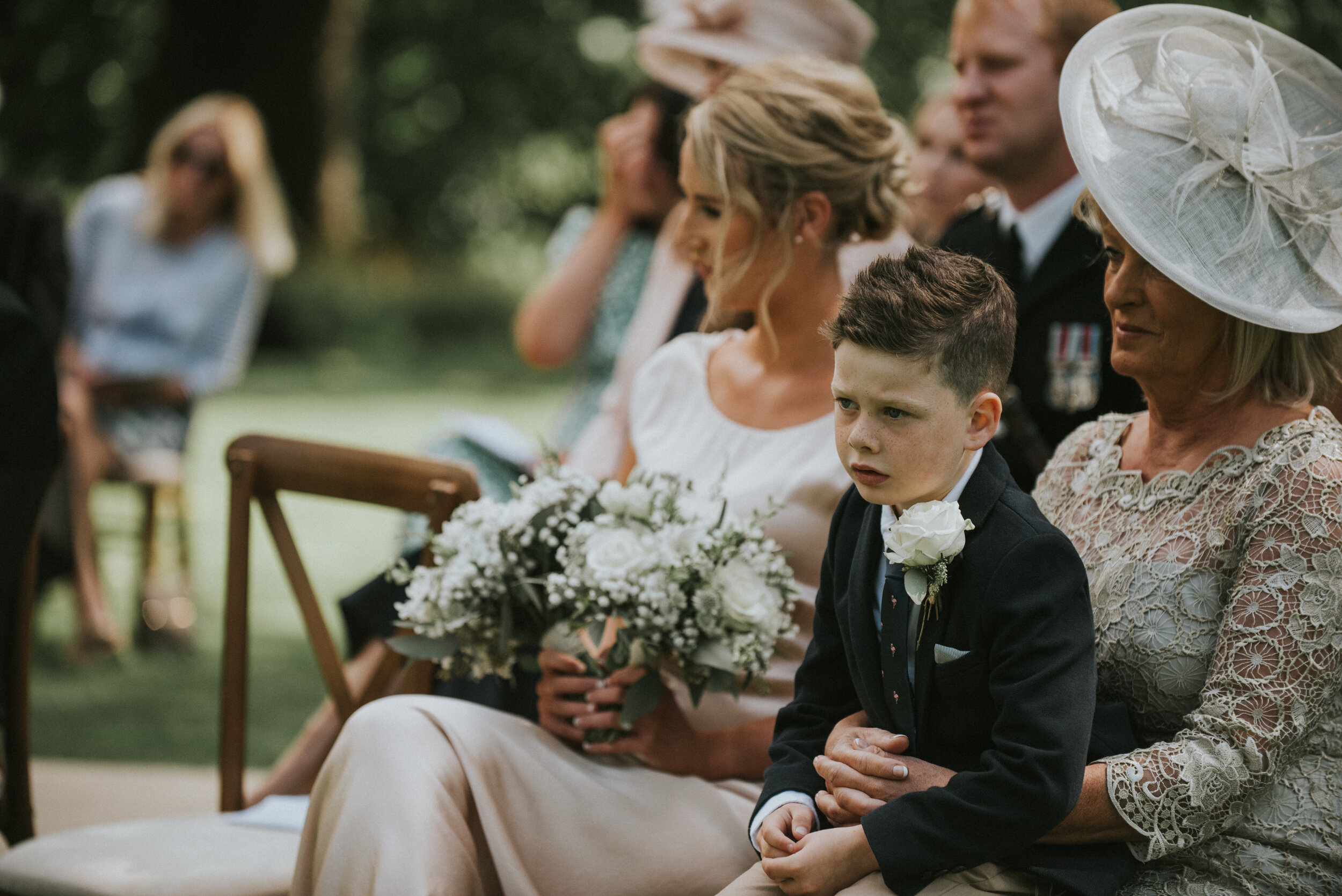 A young boy and an older woman at a wedding ceremony, with the boy in a suit and the woman wearing a large white hat and lace dress, seated outdoors with other guests around them.