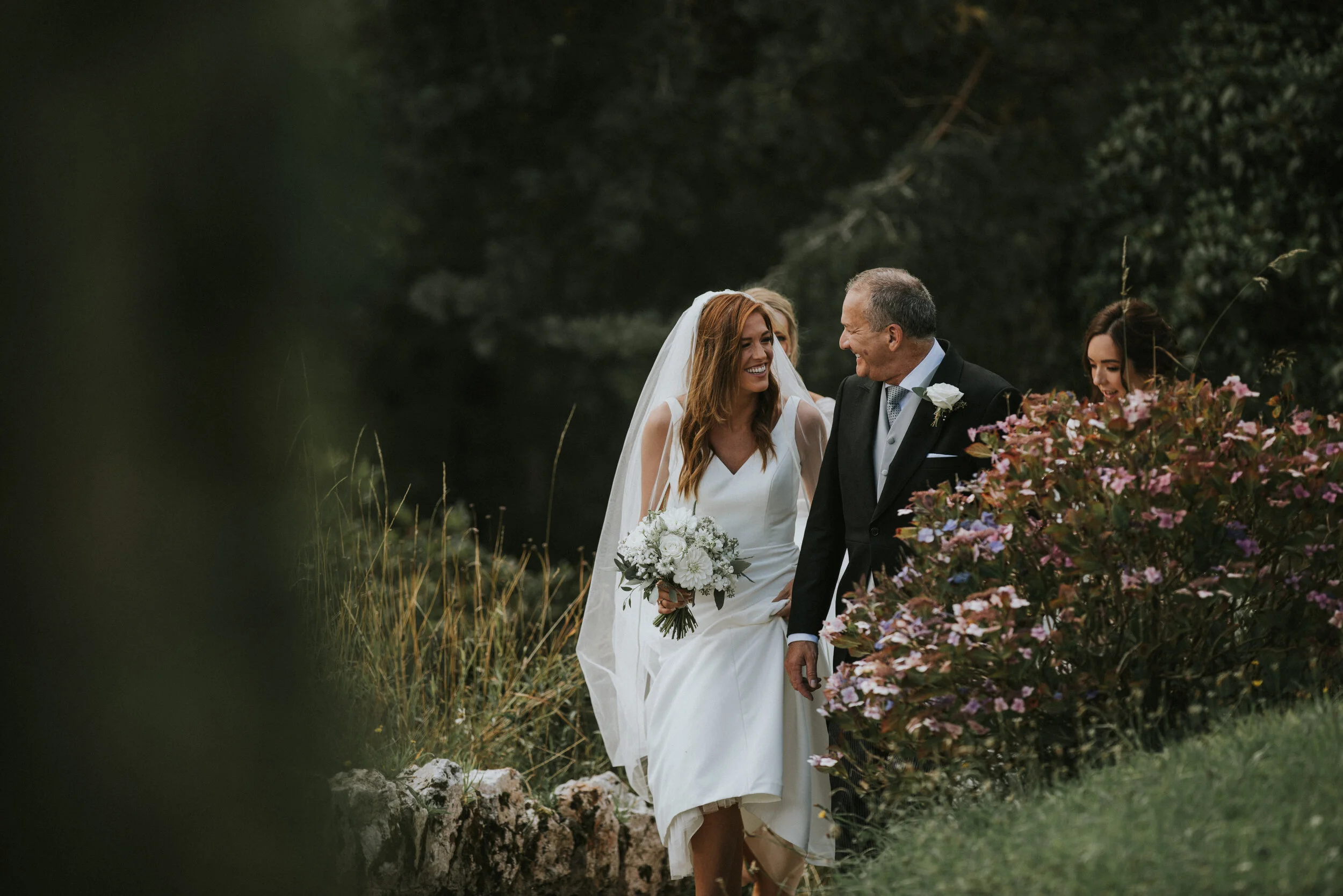 Bride and groom walking outdoors, smiling and holding flowers, with two women nearby, during a wedding celebration.