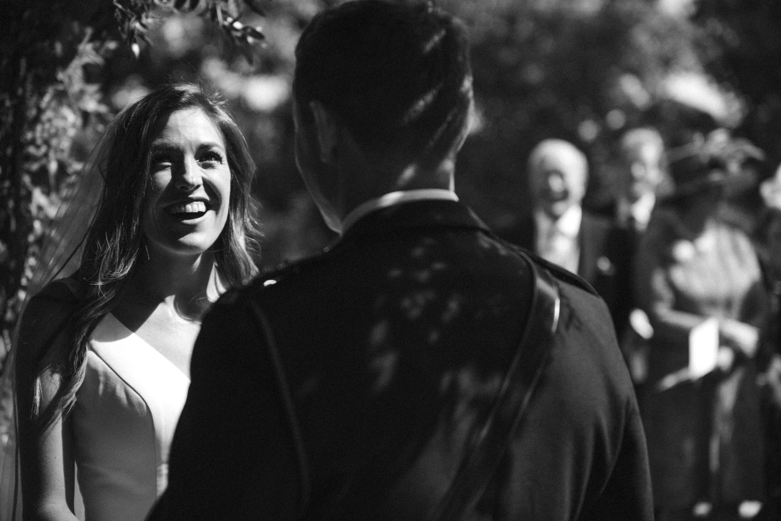 A smiling woman engaged in conversation with a man outdoors, with several people in the background during the daytime.