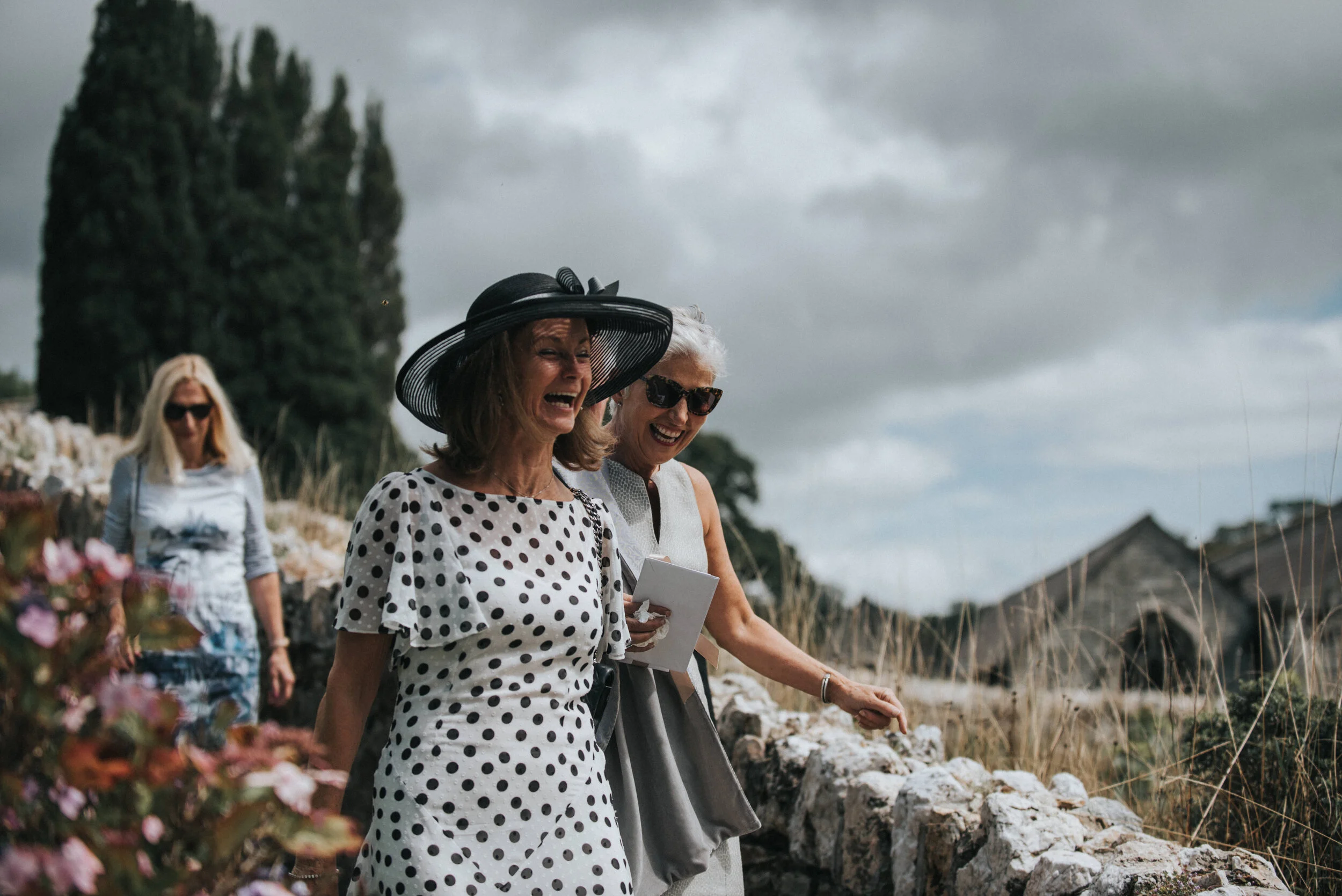 Three women walking outdoors on a cloudy day, one wearing a black hat and polka dot dress, another in sunglasses and sleeveless dress, and the third in sunglasses and a striped shirt, smiling and enjoying their walk near a stone wall with greenery an