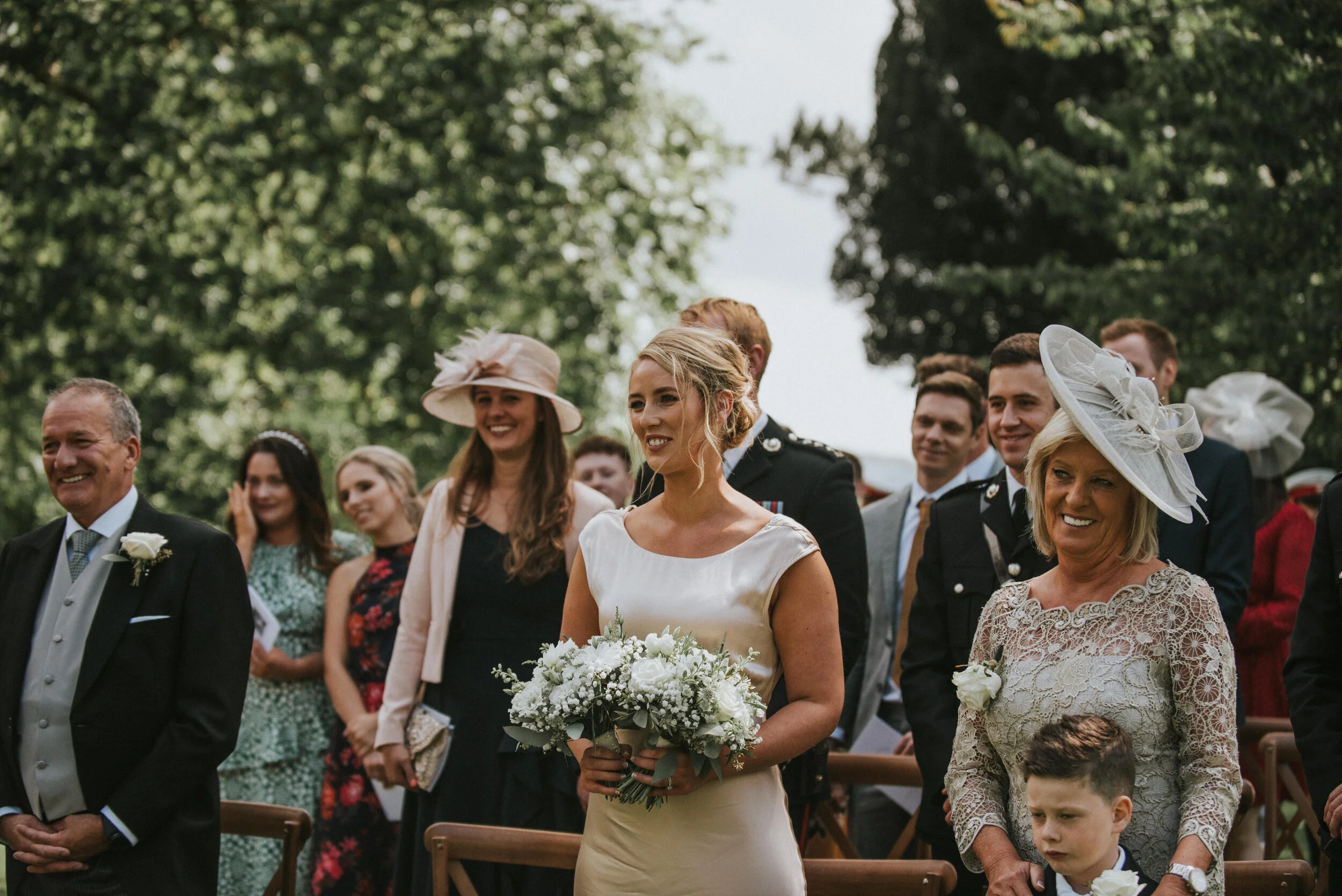 Bride holding a bouquet, smiling, during an outdoor wedding ceremony, surrounded by joyful guests dressed in formal attire.