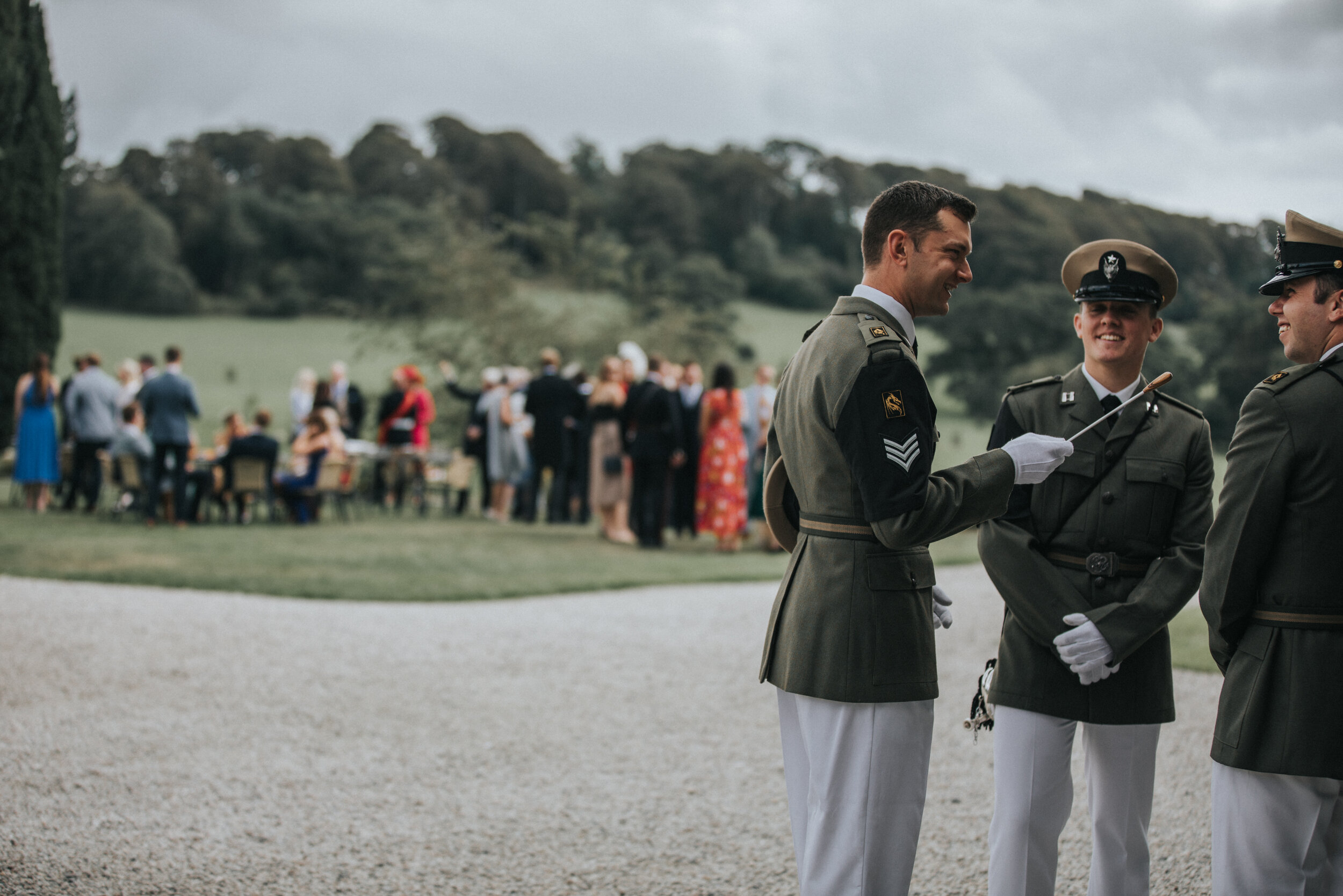 Military officer in dress uniform talking to two other officers in uniform at an outdoor social event, with a gathering of people in the background.